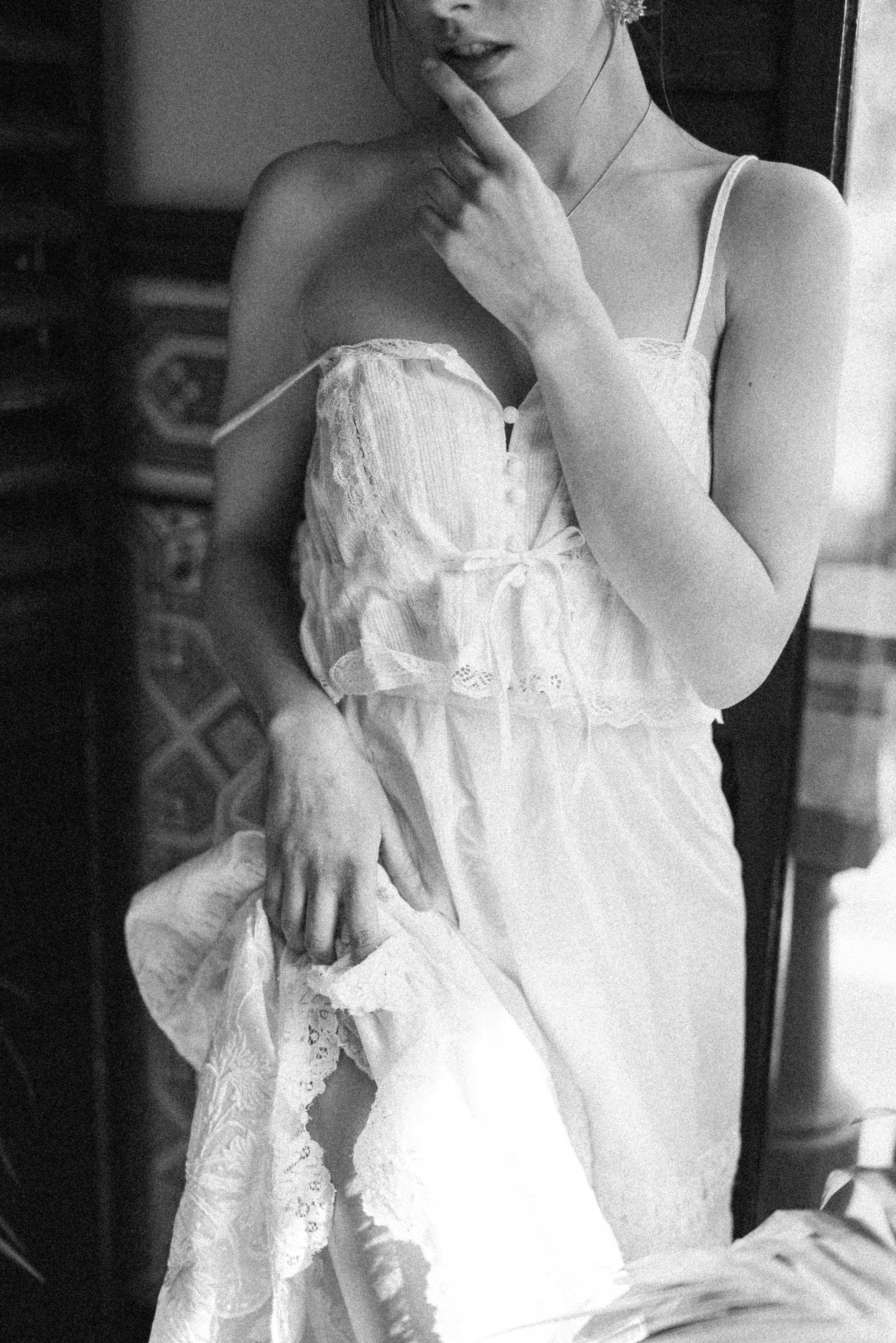 A woman wearing a lace and satin nightgown, with her finger on her lips, standing in front of a mirror in a domestic setting.