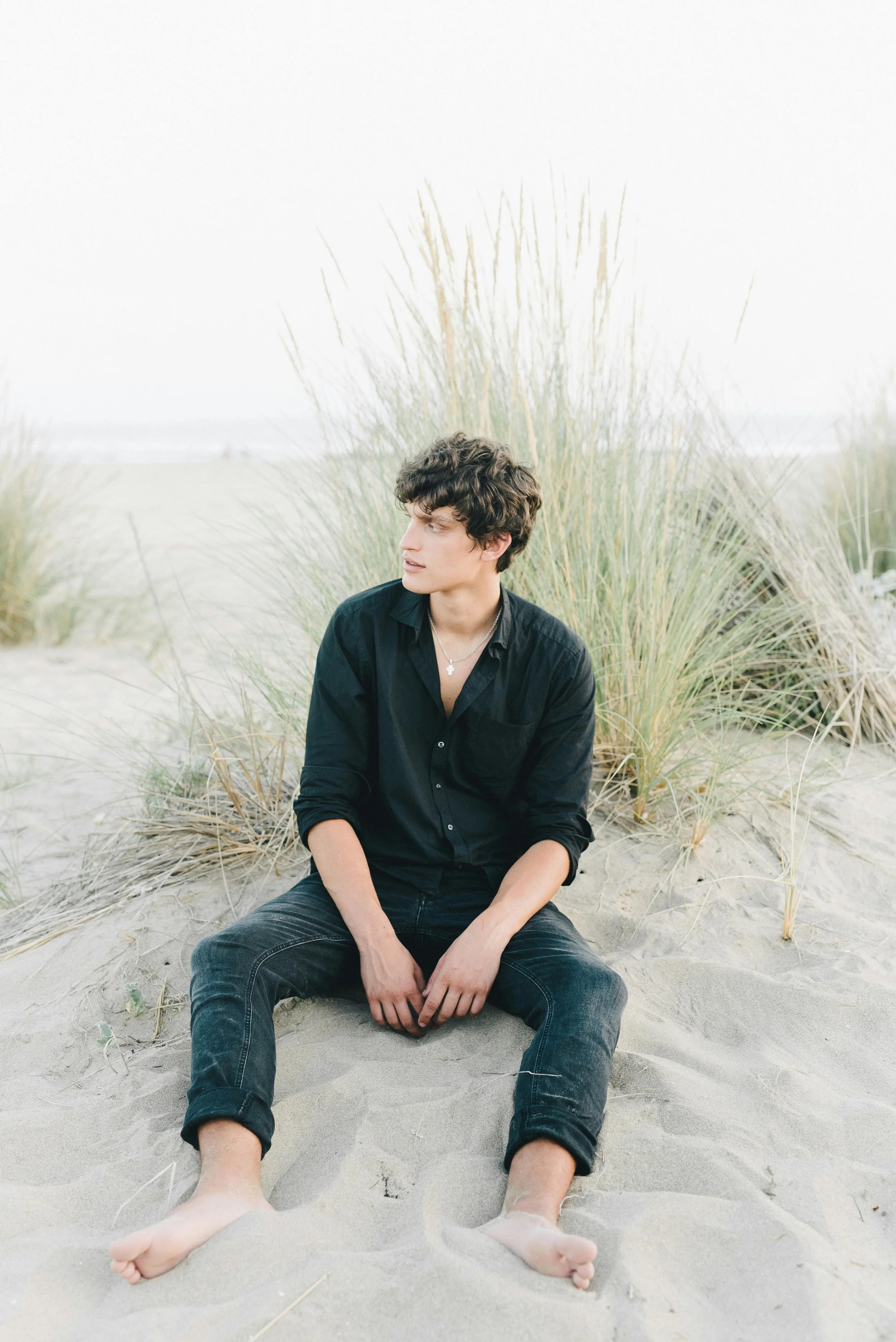 A young man with dark curly hair wearing a black shirt and black jeans sitting on sandy beach with tall grass in the background.