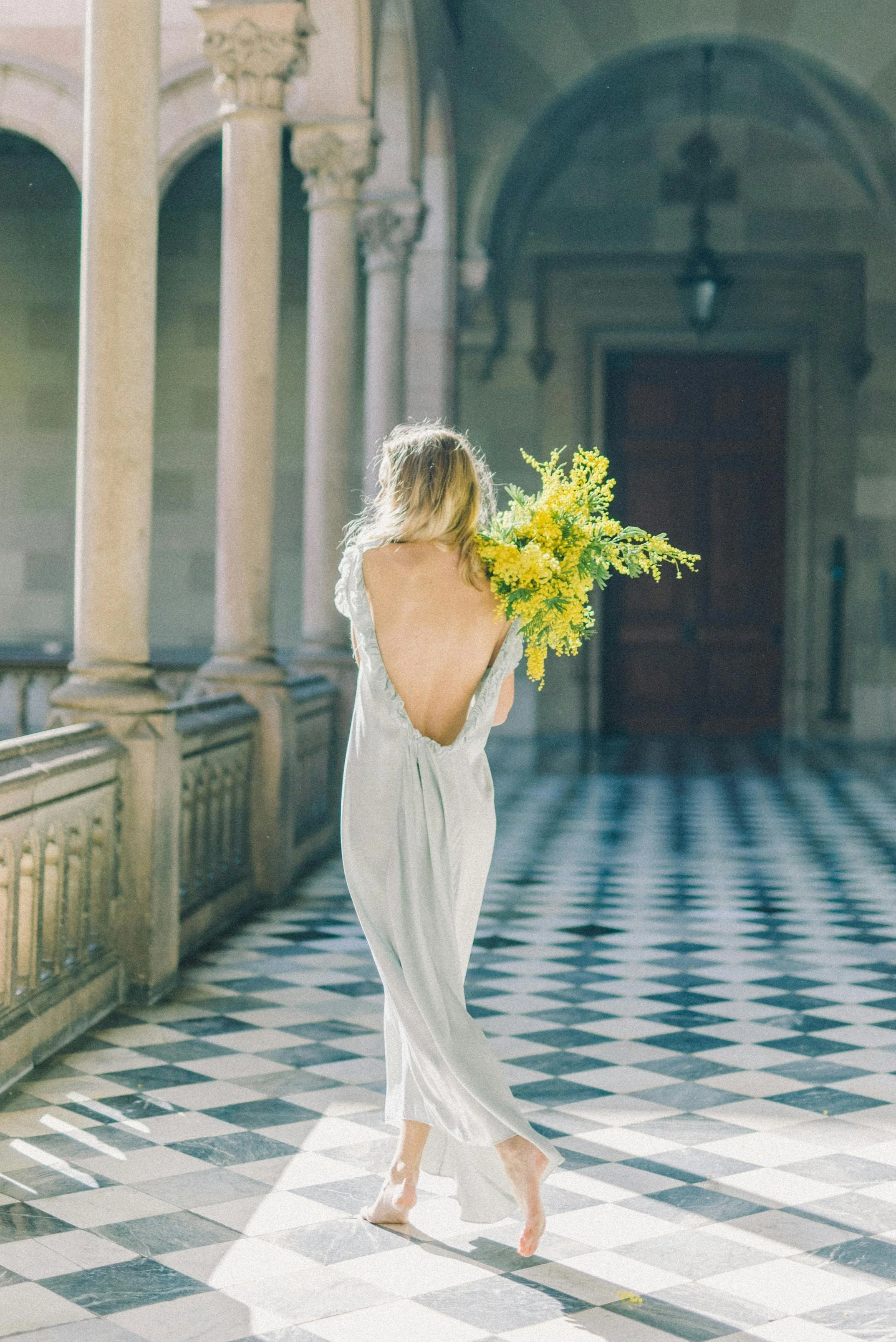 Woman in a backless, white satin dress walking barefoot through a sunlit, ornate corridor with black and white checkered floor, holding a large bouquet of yellow flowers.