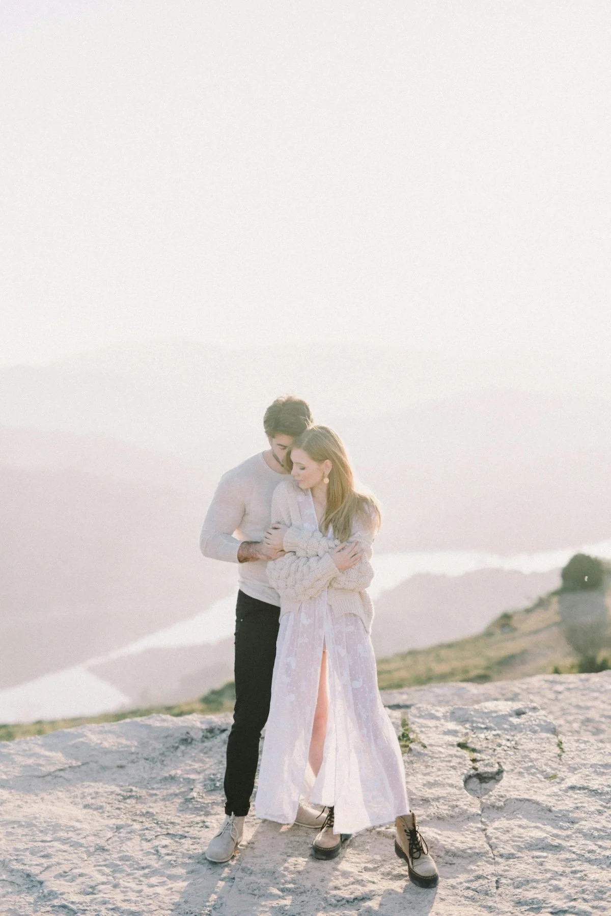 A young couple standing together outdoors on a rocky surface with hills in the background, embracing affectionately during sunset or sunrise.