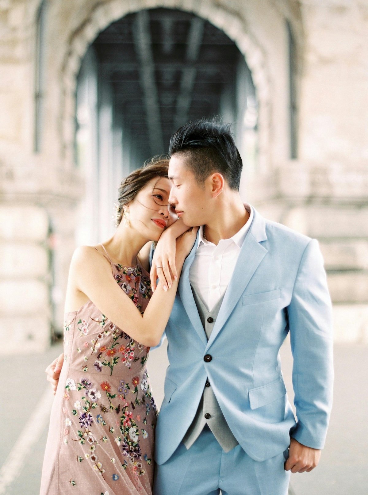 A romantic couple with Asian features standing close together under a bridge, with the woman resting her hand on the man's shoulder, both with eyes closed and foreheads touching, shared intimate moment.