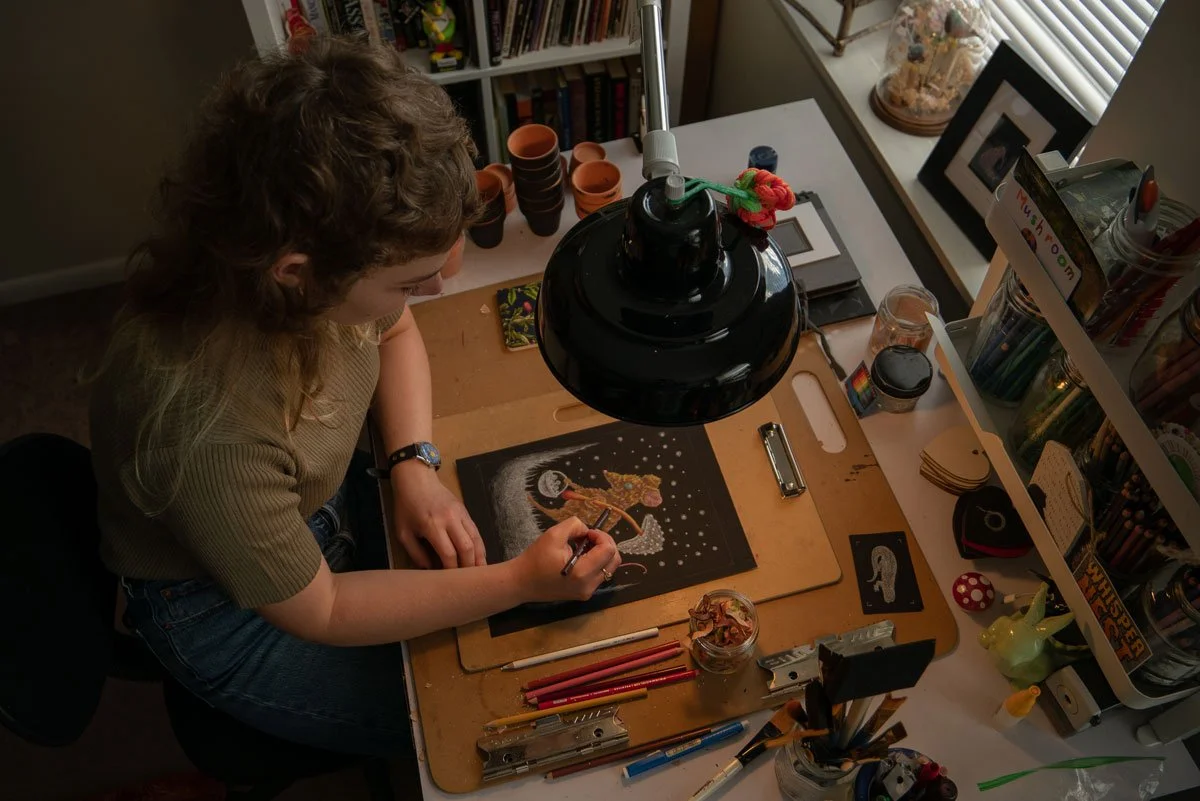A woman is working on a detailed artwork of a unicorn on a black background, at a cluttered desk with art supplies, a lamp, and various decorative items.