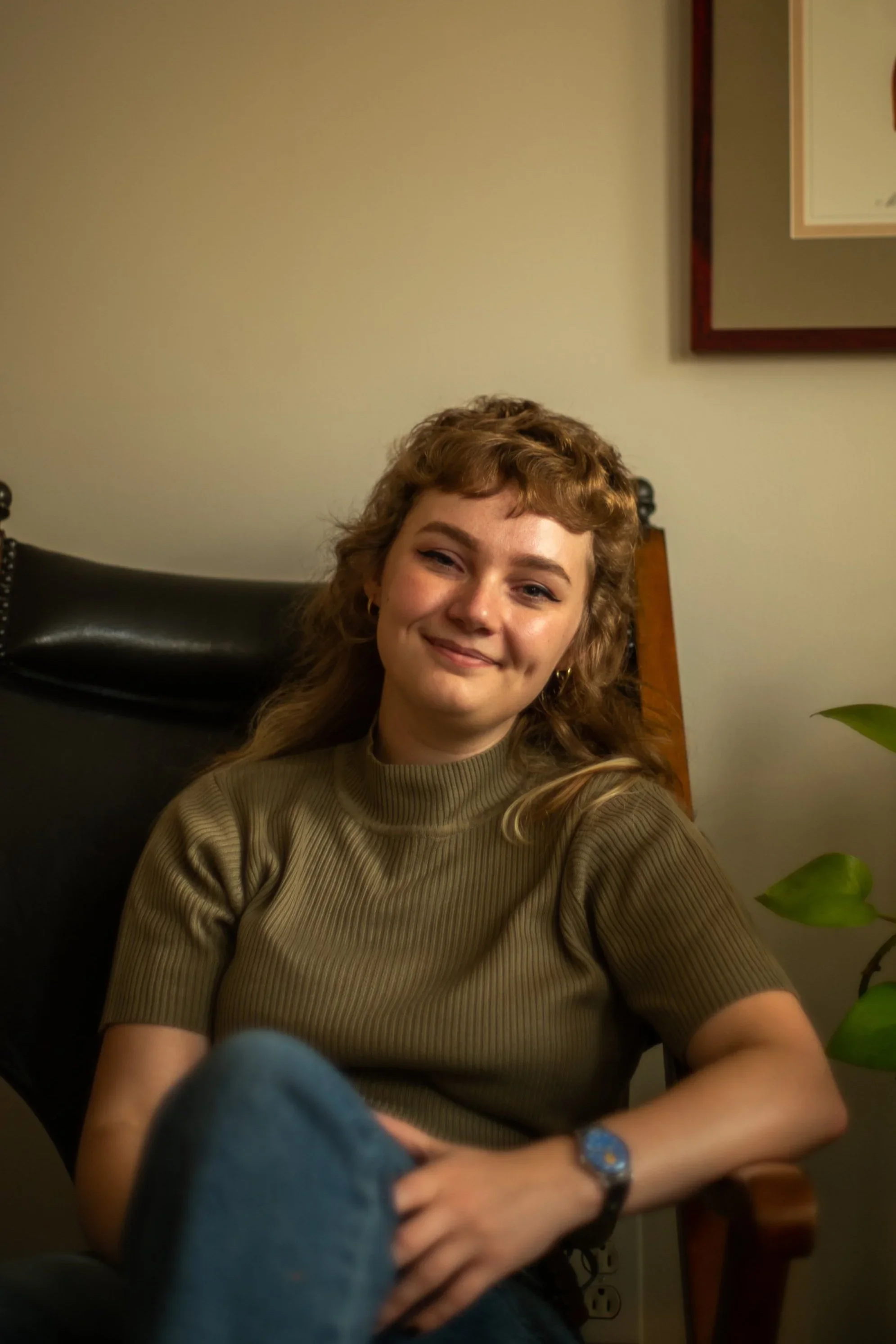 Young woman with curly hair smiling, sitting relaxed in a chair in a room with beige walls, a framed picture on the wall, and a green plant nearby.