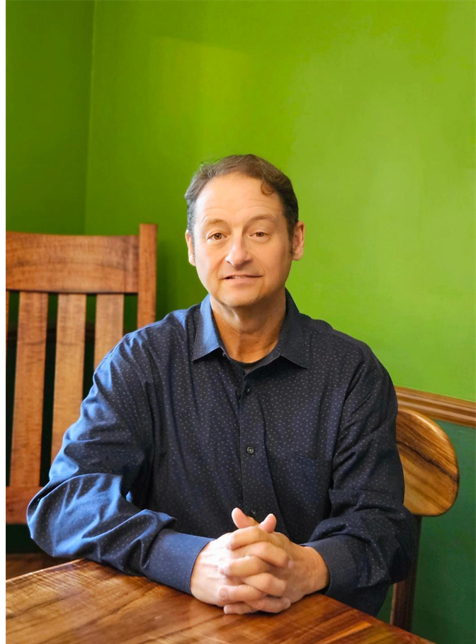 A man sitting at a wooden table with hands clasped, wearing a dark blue button-up shirt, in a room with a green wall and a wooden chair in the background.