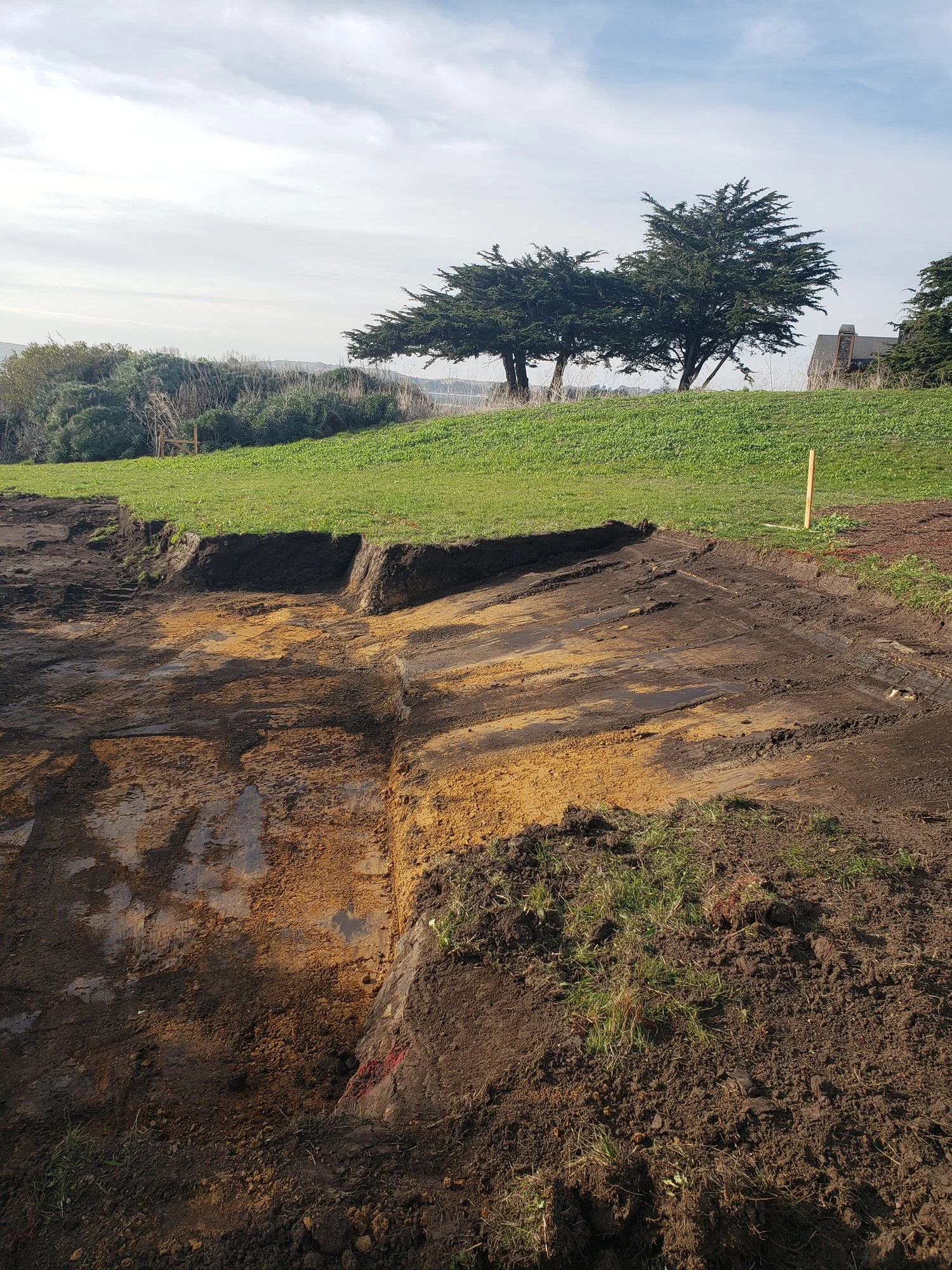 Excavation site with exposed soil and a partially excavated trench in a grassy area, with trees and a house in the background under a partly cloudy sky.