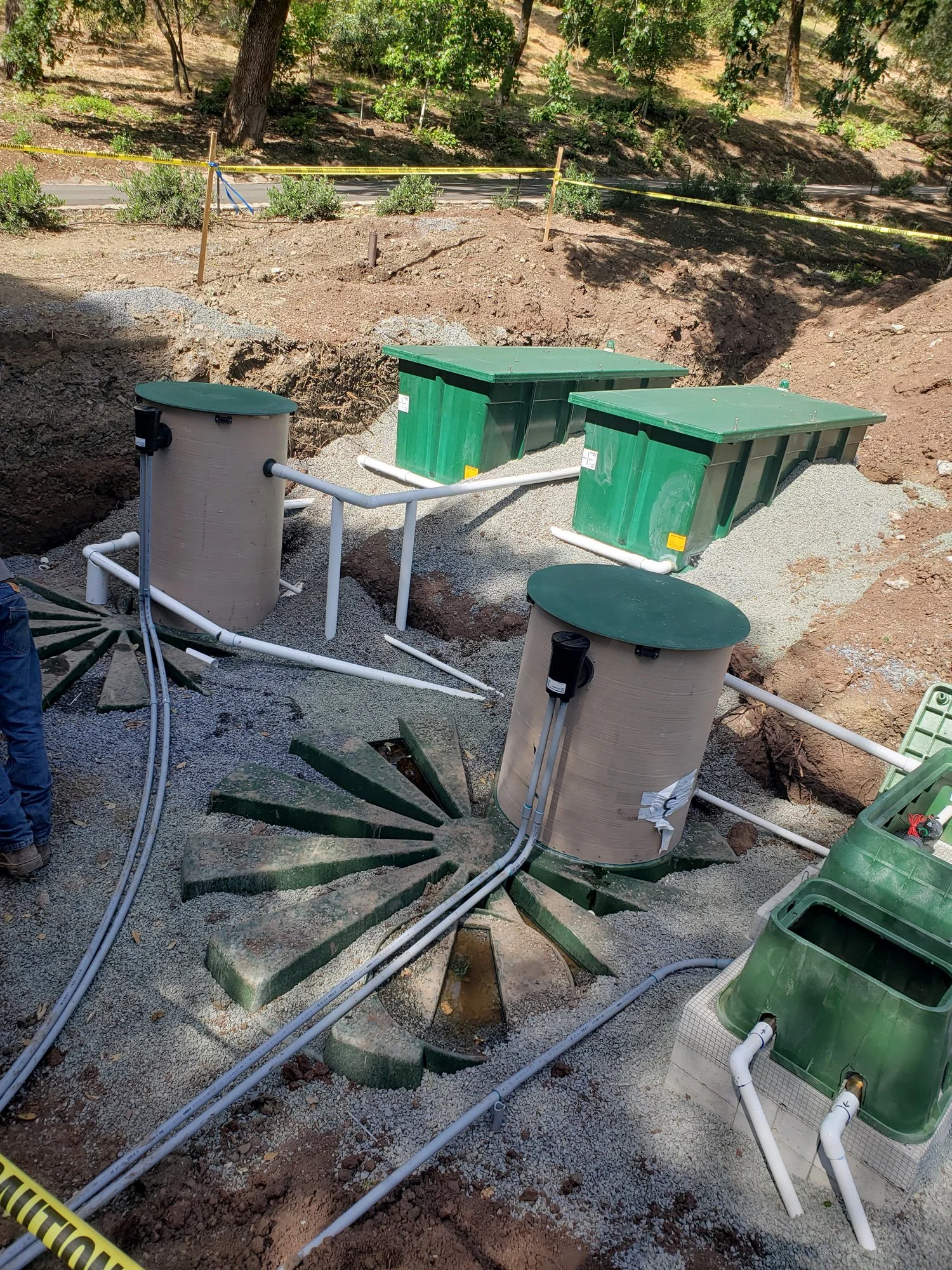 Underground utility infrastructure with green and beige tanks, connected by white pipes, situated in a construction site.