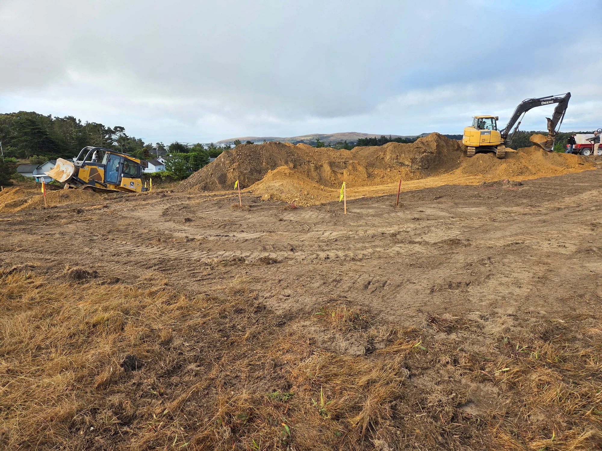 Construction site with excavators moving dirt, with a large pile of soil, and a cloudy sky overhead.