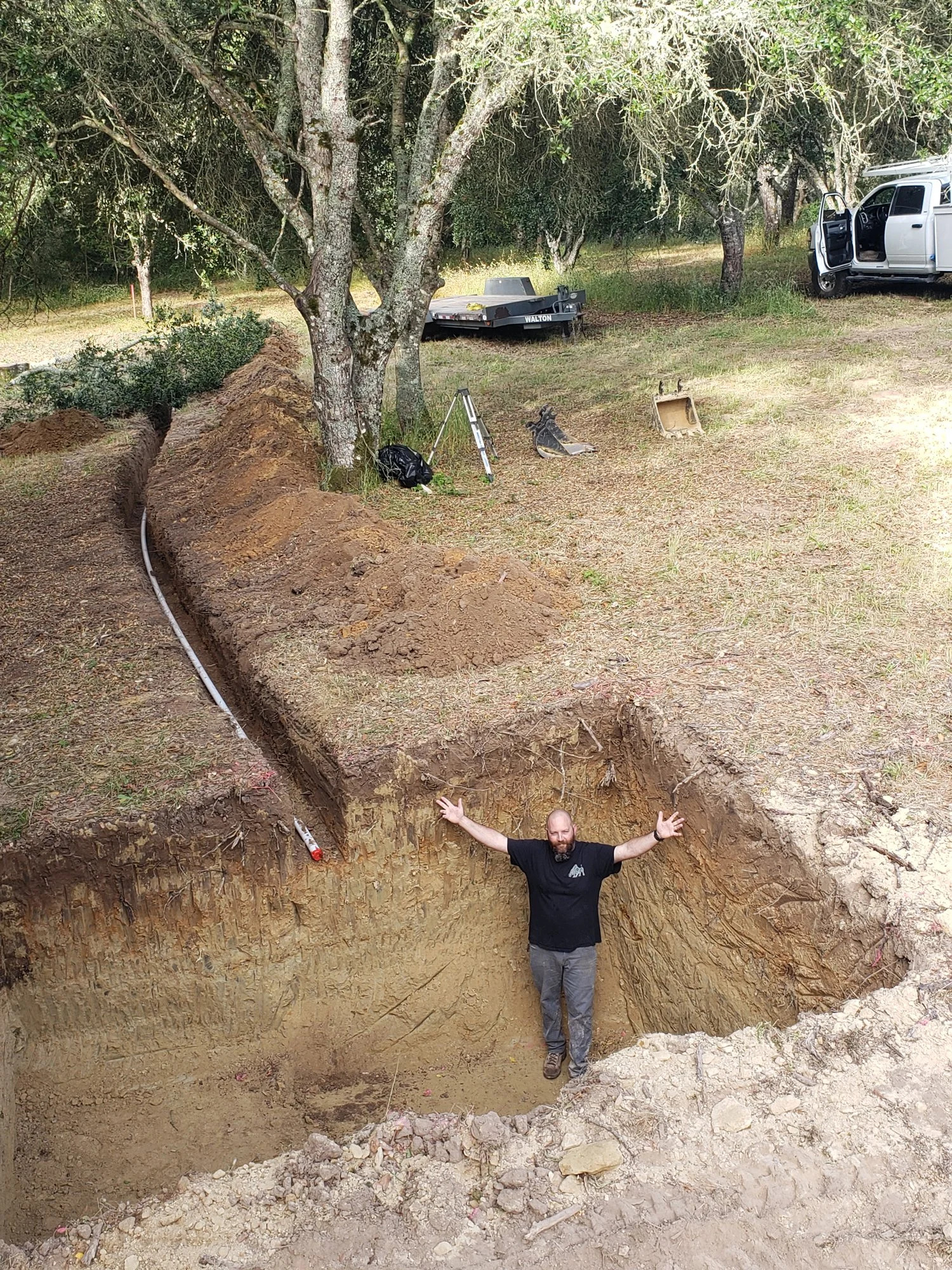A man standing inside a large excavation site with arms outstretched, surrounded by dirt and trees, with construction equipment and a vehicle nearby.