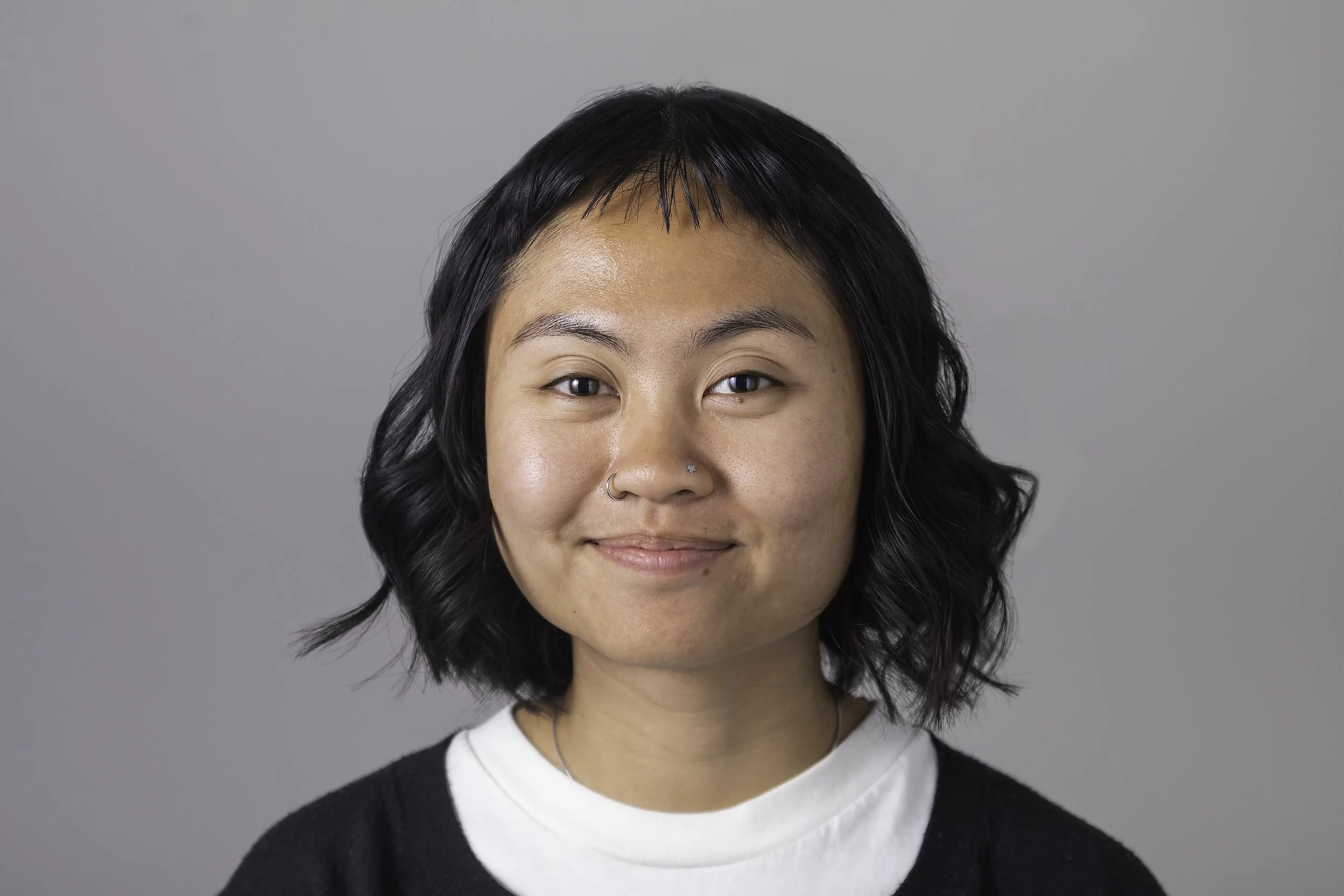Portrait of a young woman with short, wavy black hair, wearing a nose ring and a black and white top, smiling against a plain gray background.
