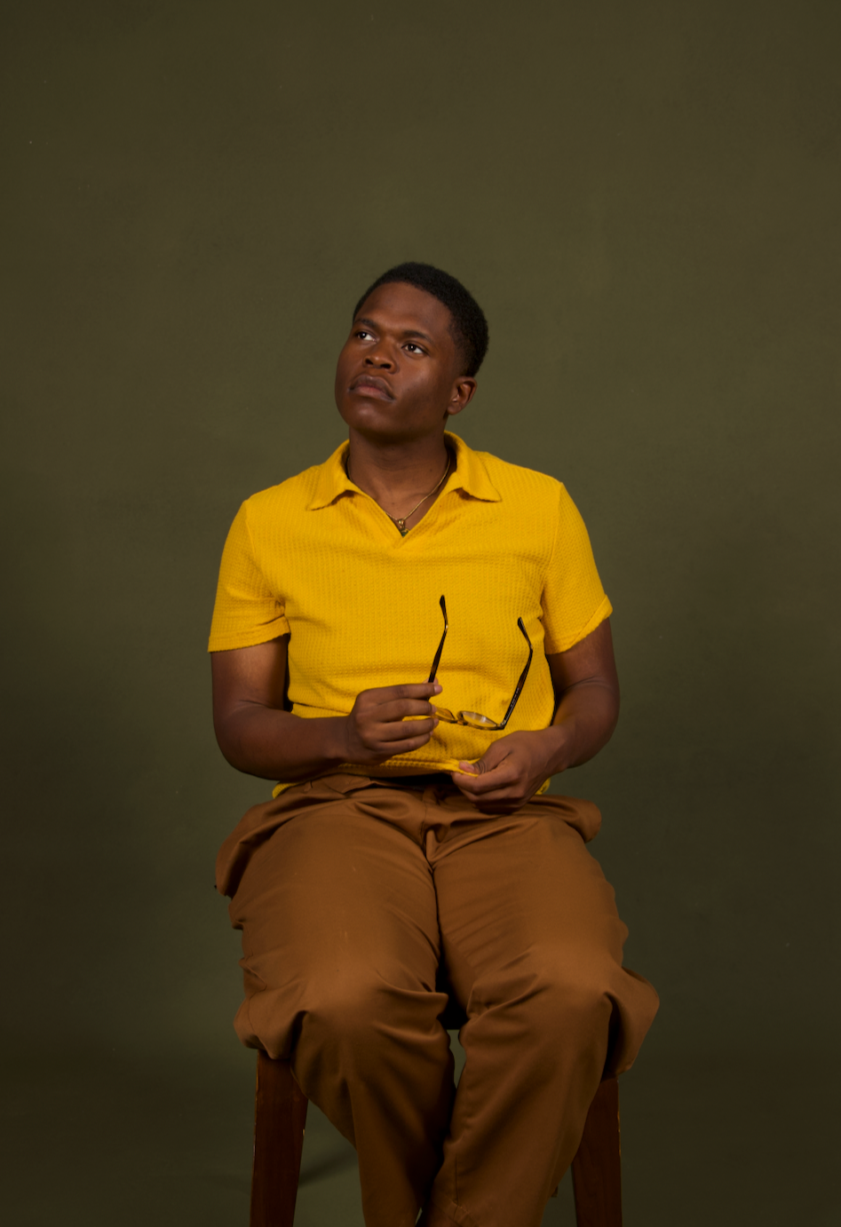 A young man wearing a yellow shirt and brown pants is sitting on a wooden stool against a plain olive-green background. He is holding glasses in one hand and looking thoughtfully upward.