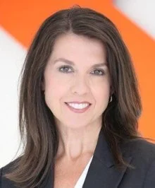 A man with wavy dark hair, light facial hair, wearing glasses and a nose ring, looking slightly to the side with a soft smile, sitting in a bright room with a blurred background and a plant.