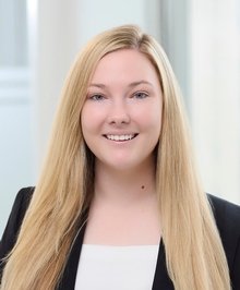 Young woman with long blonde hair smiling, wearing a black blazer and white top, in an indoor setting with bright natural light.