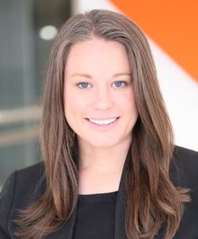 Portrait of a woman with long brown hair, wearing a black top and blazer, smiling in an indoor setting.