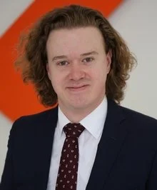 A man with shoulder-length wavy brown hair wearing a dark suit, white shirt, and a maroon polka dot tie, standing against an orange and gray background.