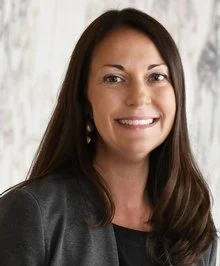 Smiling young woman with glasses and curly hair, wearing a striped shirt and gold jewelry, indoors.