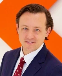 A man with curly dark hair, glasses, and a beard, wearing a dark green textured sweater, looking off to the side with a slight smile.