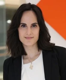Professional woman with shoulder-length dark hair wearing a black blazer and white top, standing indoors with a blurred background and an orange accent wall.
