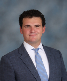 Professional headshot of a man in a suit and tie smiling against a gray background.