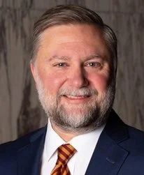 A woman with straight black hair smiling outdoors surrounded by green plants.