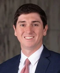 Smiling Asian woman with shoulder-length black hair wearing a blue top, standing among green foliage.