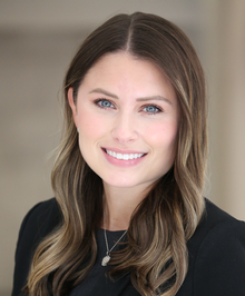 A woman with long brown hair wearing a black top and smiling, in a well-lit indoor setting.