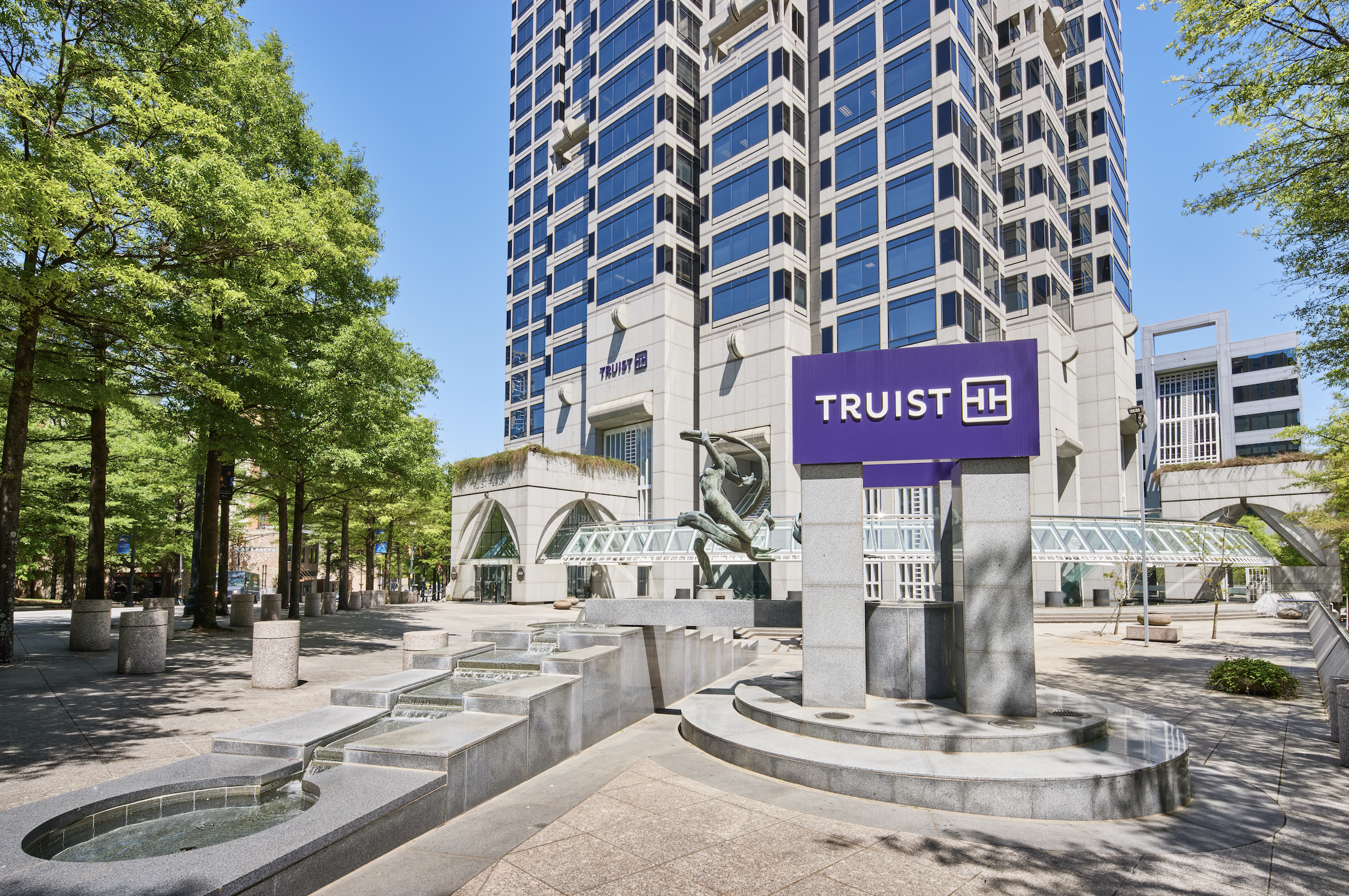 A city scene with a tall modern office building, trees lining the street, a purple sign with 'TRUIST', a fountain with statues, and a sculpture of a human figure climbing.