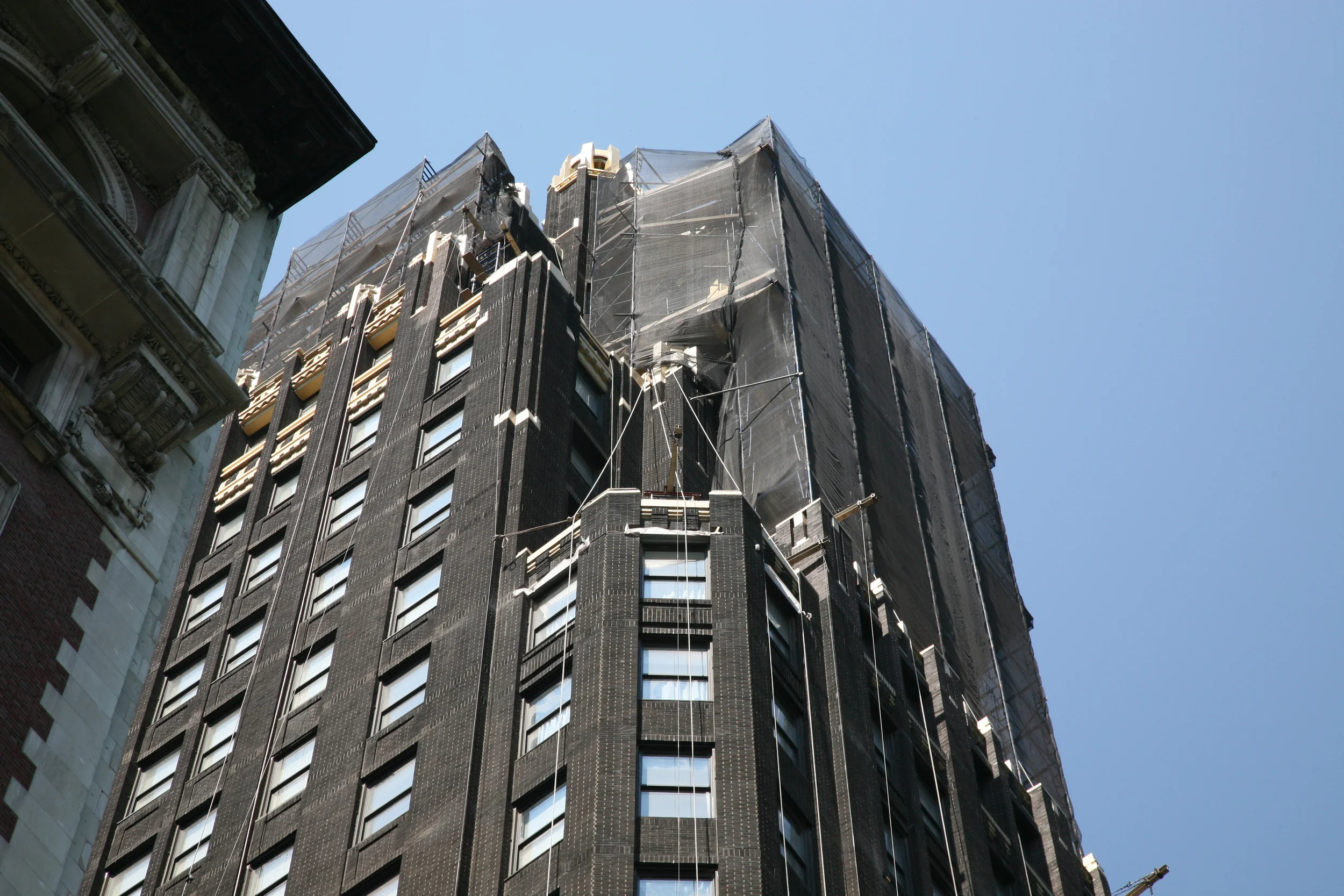 A tall building under renovation, covered with scaffolding and black protective netting, against a clear blue sky.