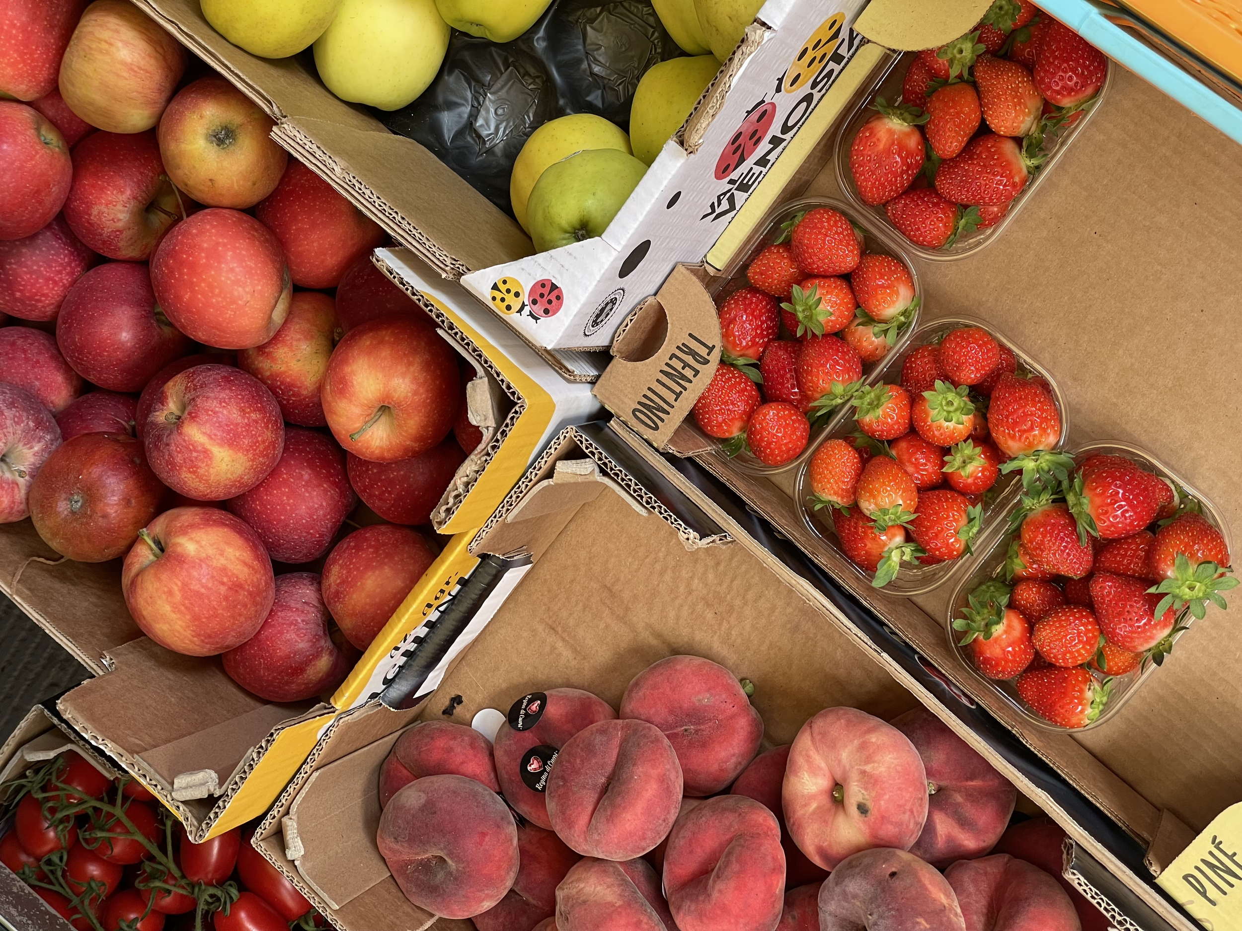 A variety of fresh fruits arranged in open boxes, including red apples, peaches, strawberries in clear containers, and cherry tomatoes.