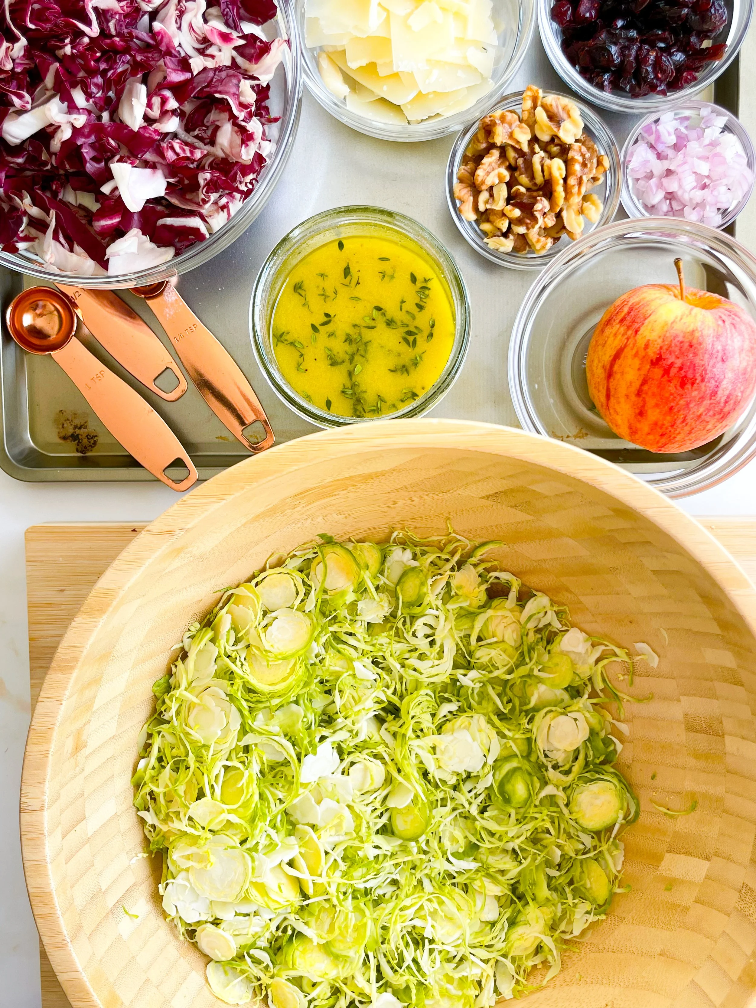 Chopped radicchio in a bowl, shredded cheese in a bowl, dried cranberries in a bowl, chopped shallots in a bowl, walnut halves in a bowl, whole apple in a bowl, fresh chopped fennel in a large wooden bowl, small bowl of vinaigrette with herbs, copper