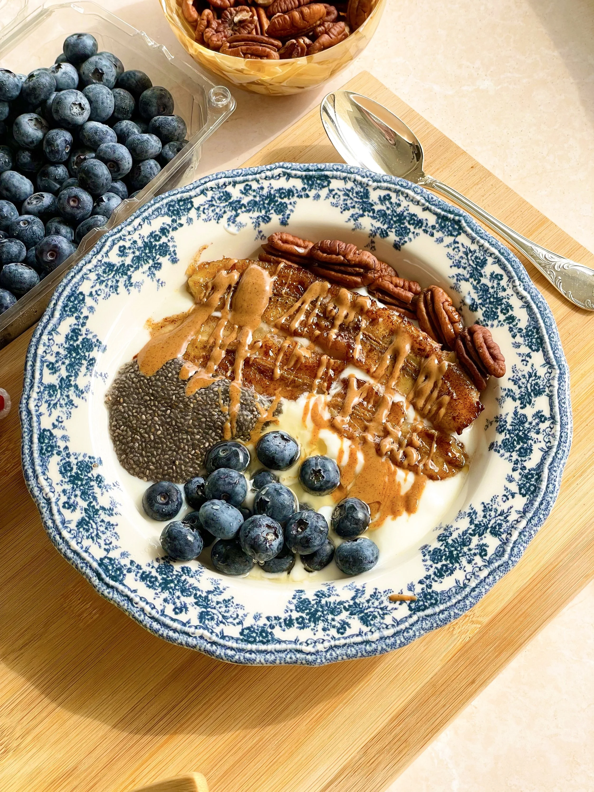 A bowl of yogurt topped with blueberries, pecans, chia seeds, and caramel sauce, placed on a wooden board.