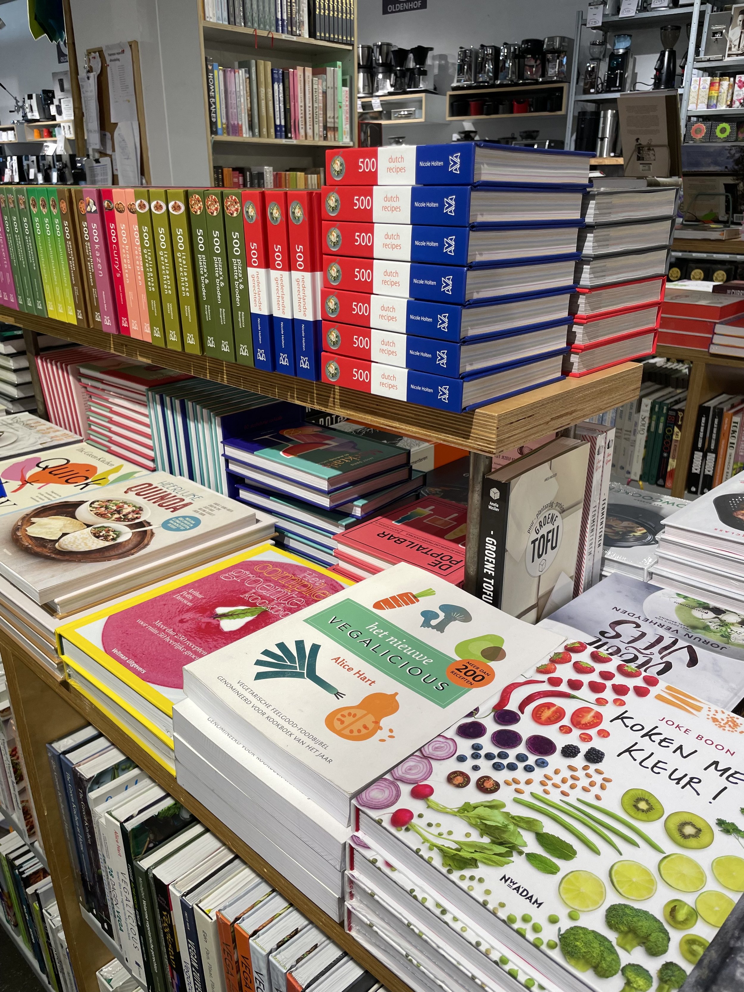 A display of cookbooks on a wooden table and shelf in a bookstore, including books about healthy vegan cooking, Dutch recipes, and various illustrated cookbooks, with shelves of books and coffee brewing equipment in the background.