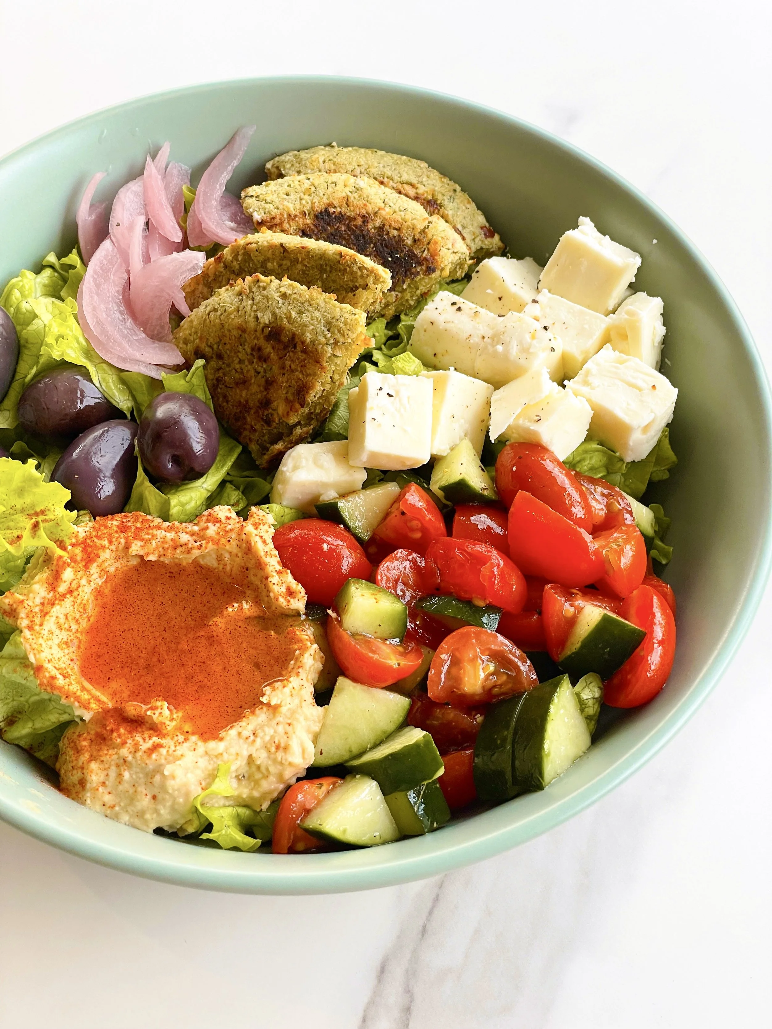 A bowl of fresh Mediterranean salad with cherry tomatoes, cucumbers, feta cheese, black olives, pickled onions, falafel pieces, and hummus with paprika, on a white marble surface.