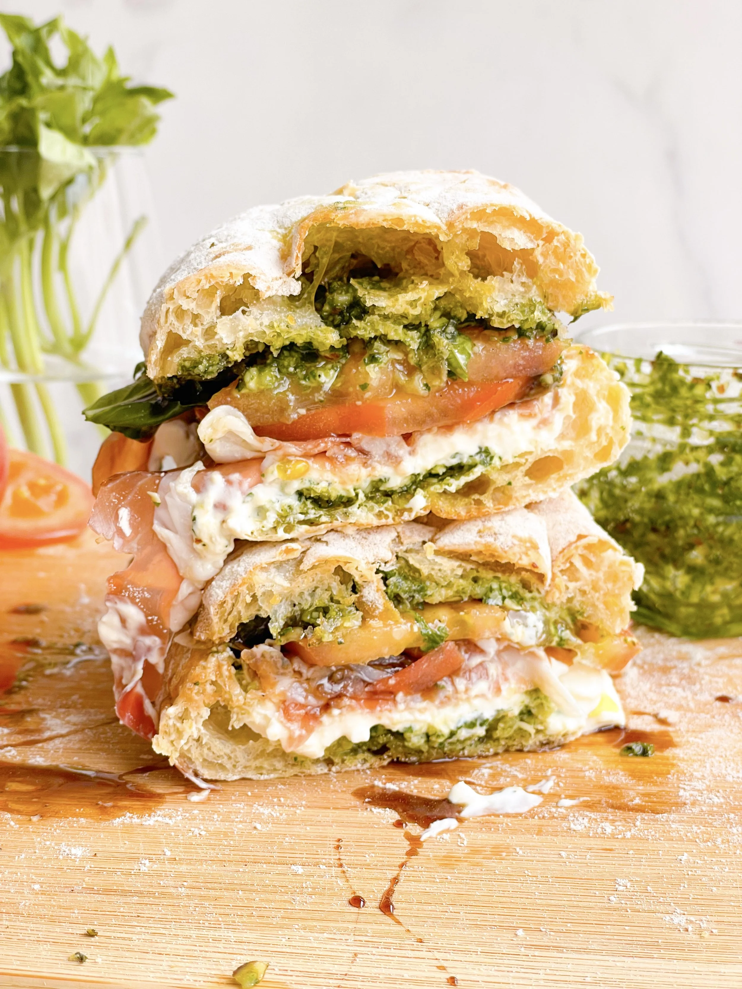 Two halves of a freshly made sandwich stacked on a wooden cutting board. The sandwich contains green pesto, sliced tomato, lettuce, cheese, and possibly ham or turkey, with some crumbs and oil on the board. In the background, a glass jar of herbs and