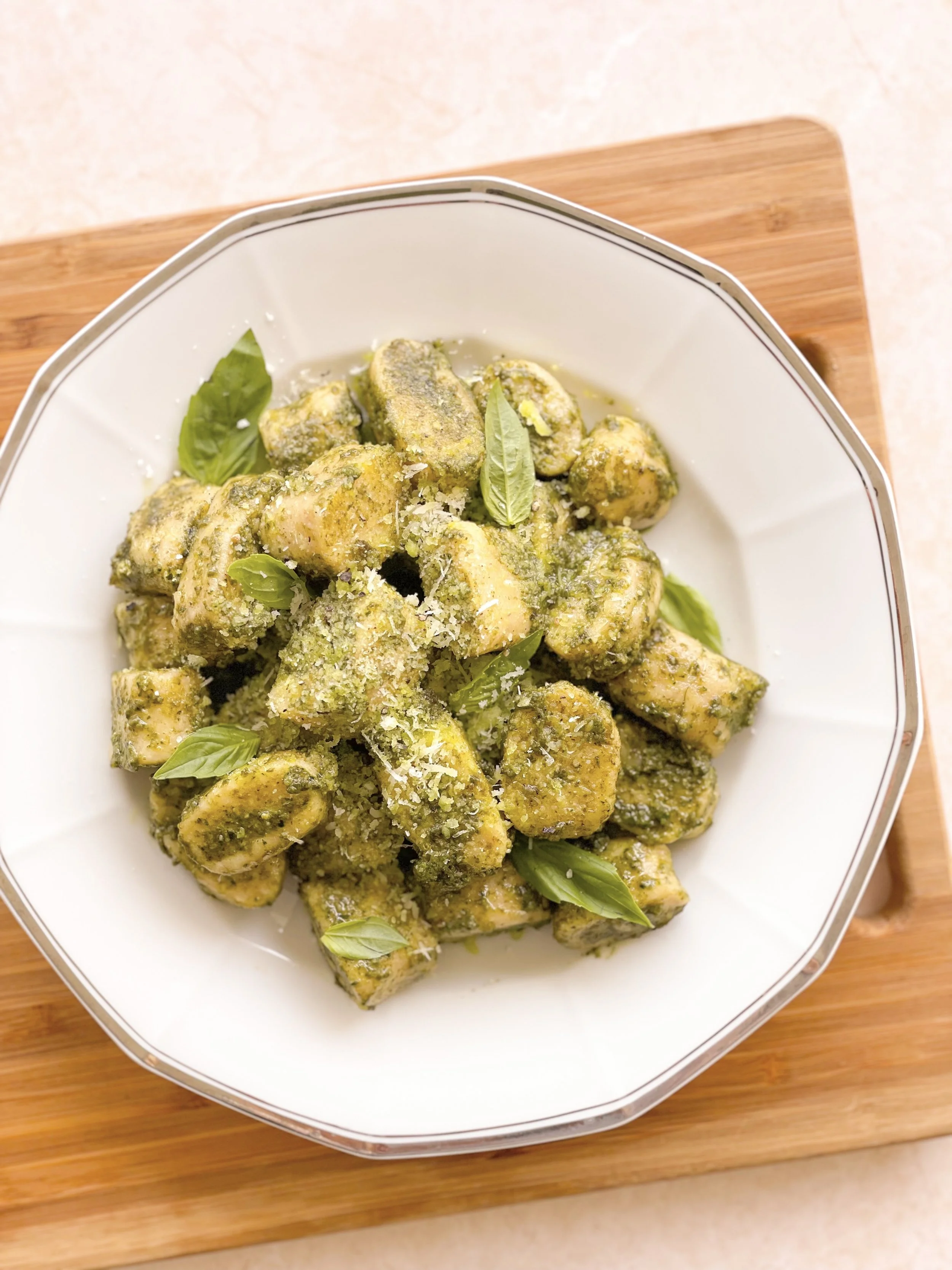 Freshly cooked gnocchi coated with green pesto sauce, garnished with basil leaves and grated cheese in a white bowl on a wooden cutting board.
