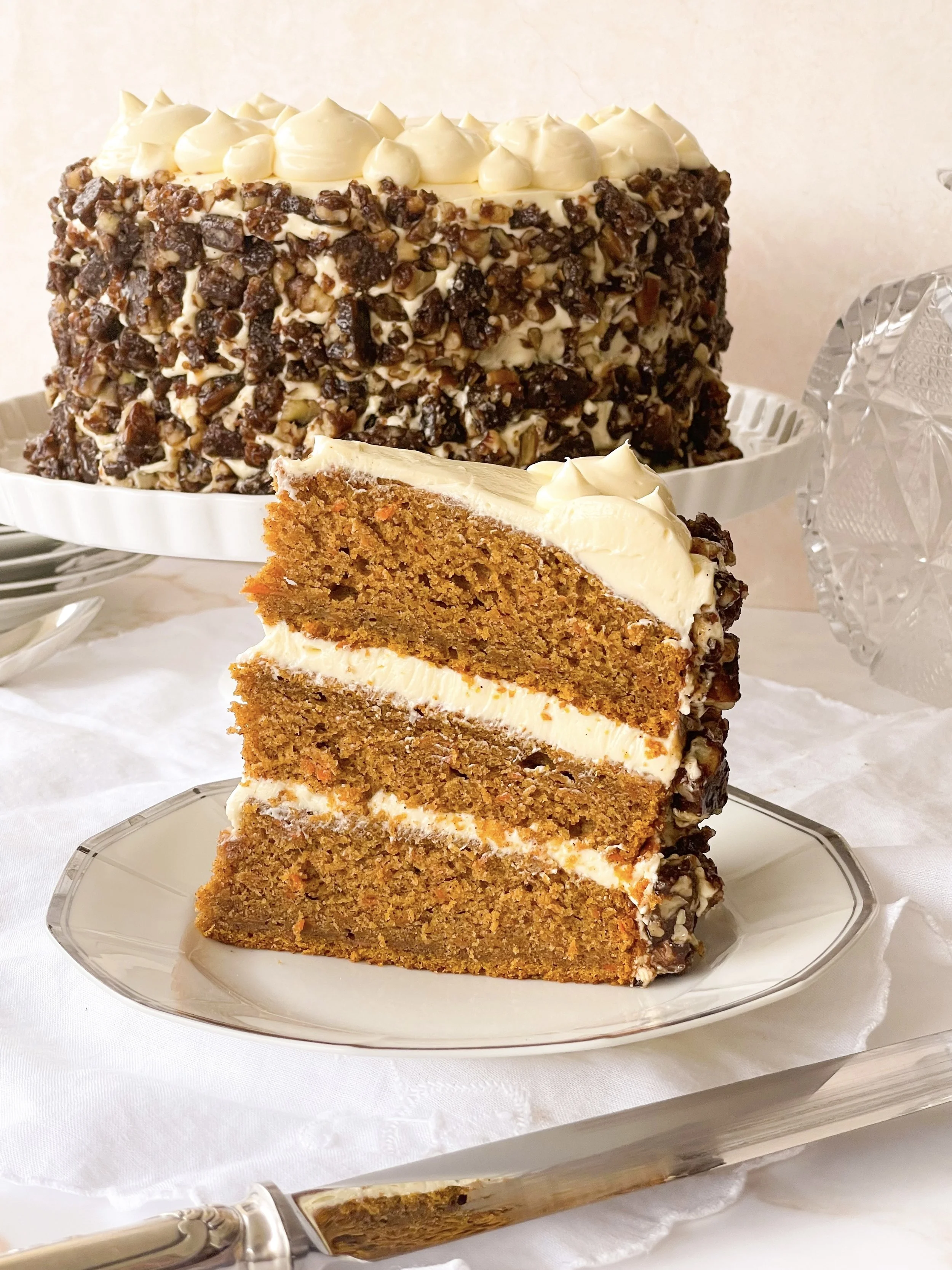 A slice of carrot cake with cream cheese frosting on a plate in front of a whole carrot cake covered in chopped nuts and cream cheese frosting.