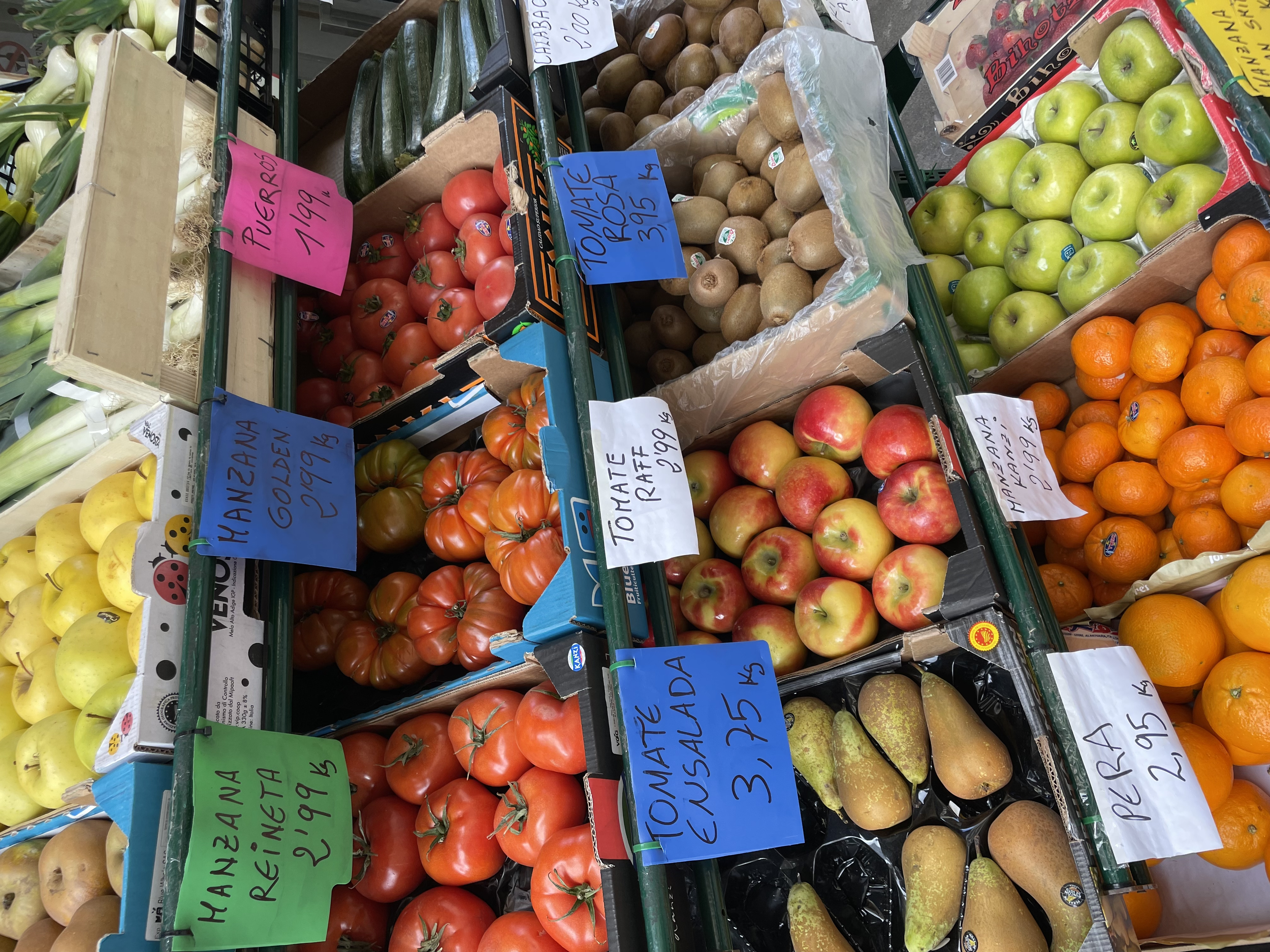 Table of fresh produce at a market, featuring tomatoes, pears, apples, oranges, and potatoes, with handwritten signs displaying prices in Spanish.