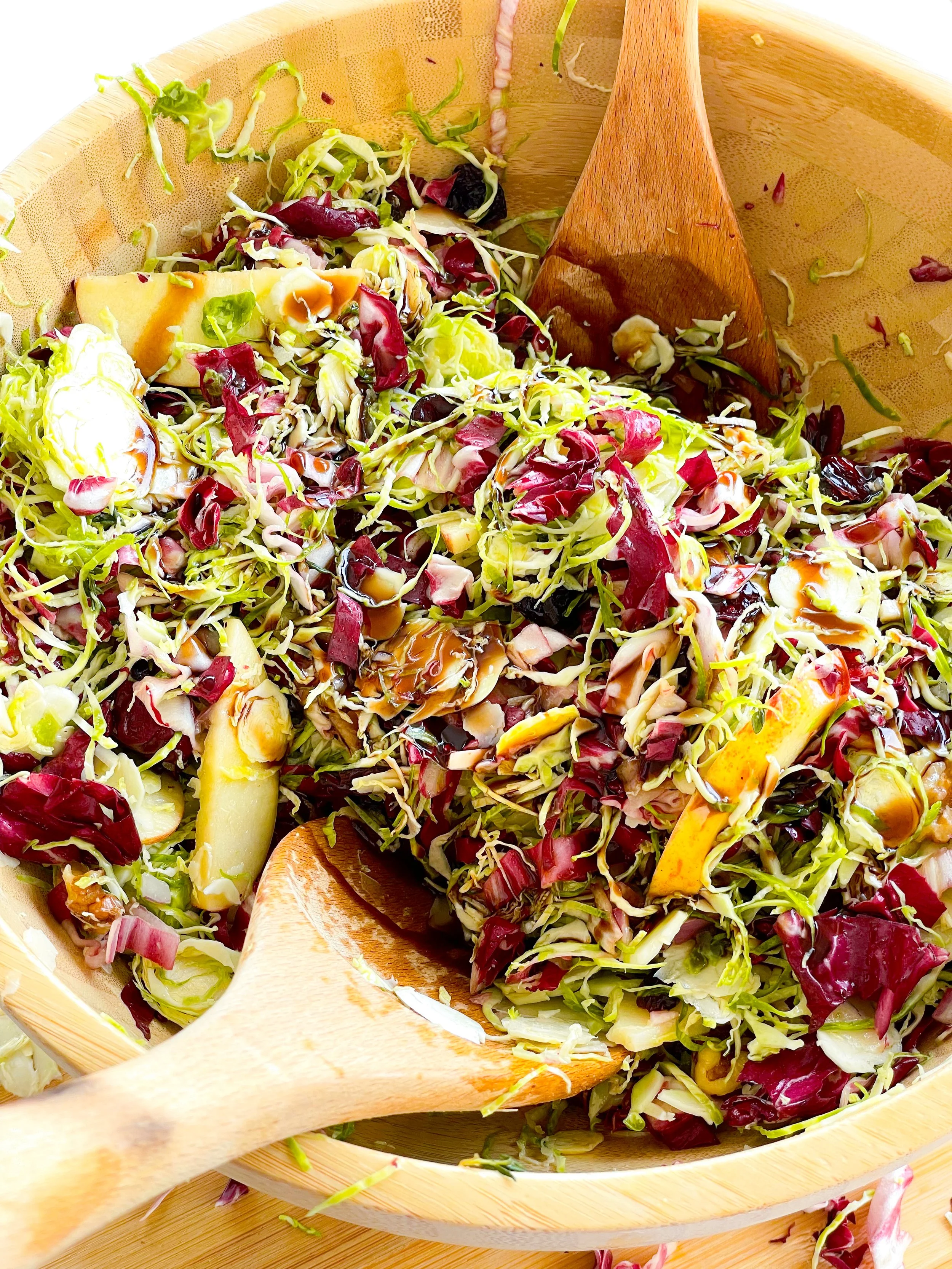 A wooden salad bowl filled with a mixture of shredded greens, radicchio, and sliced vegetables, with two wooden salad servers resting inside.