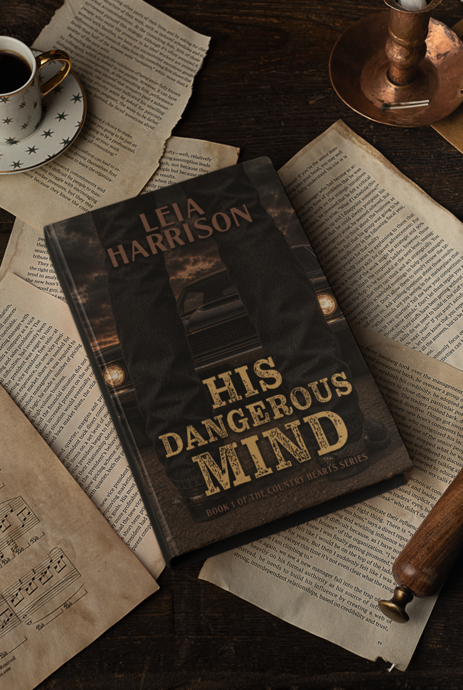 A book titled 'His Dangerous Mind' by Leia Harrison placed on open books and papers, next to a cup of coffee, a candle holder, and a wooden rolling pin on a dark wooden table.
