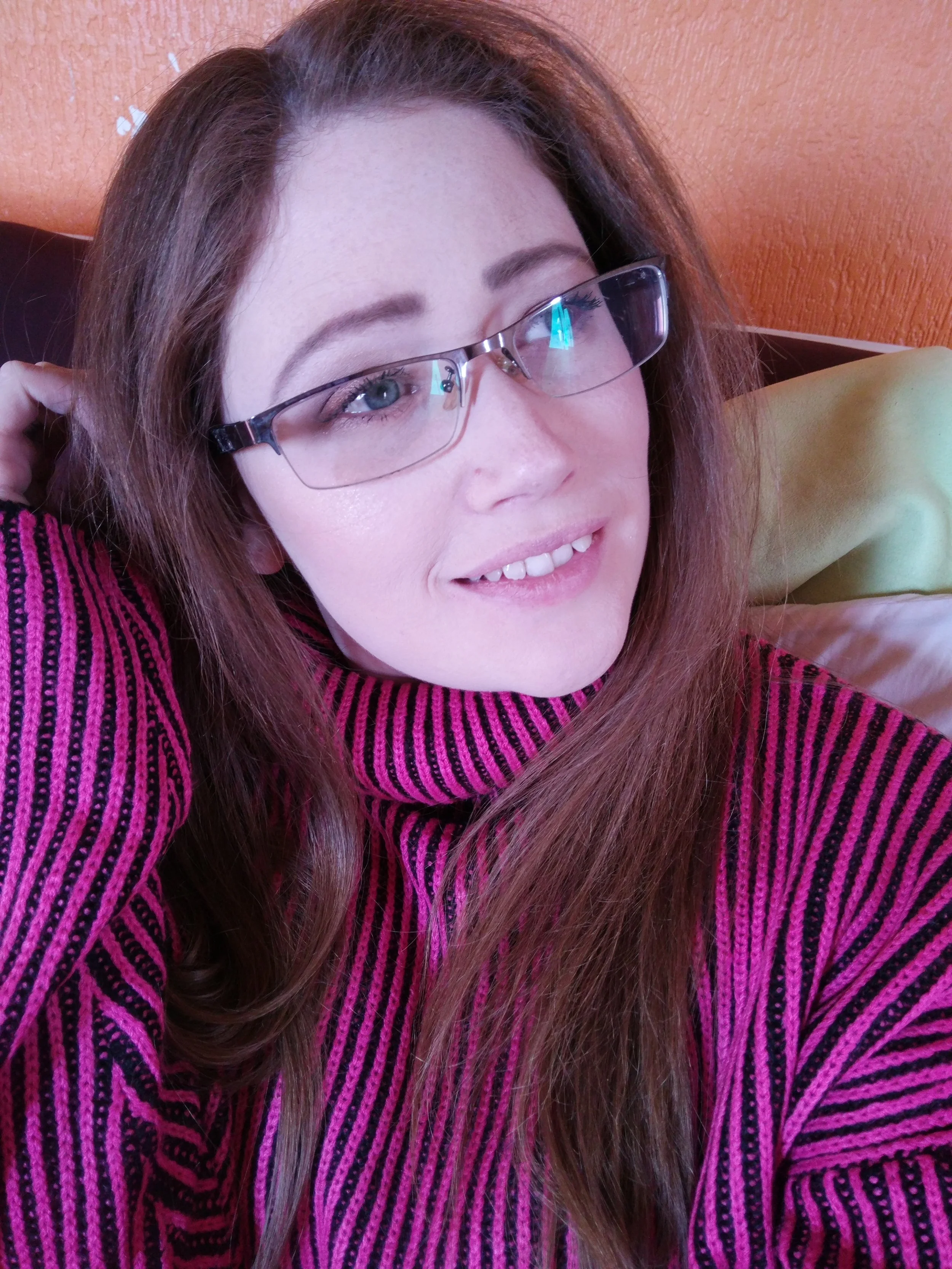 A woman with long, light brown hair and glasses smiling, wearing a pink and black striped sweater, sitting in a room with orange textured wall and light green pillow.