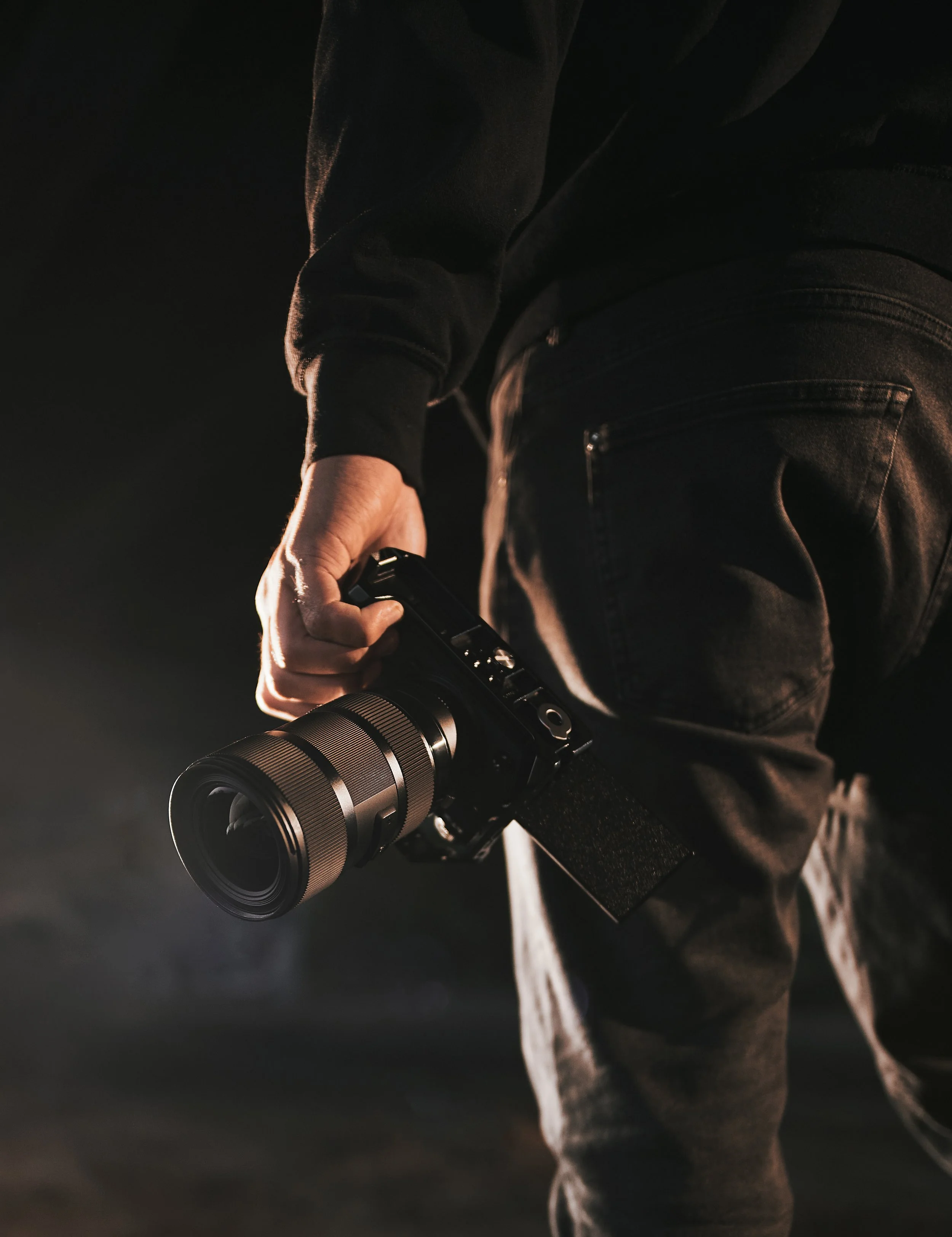 Person holding a camera with a zoom lens, wearing dark clothing and jeans, in a dark setting.