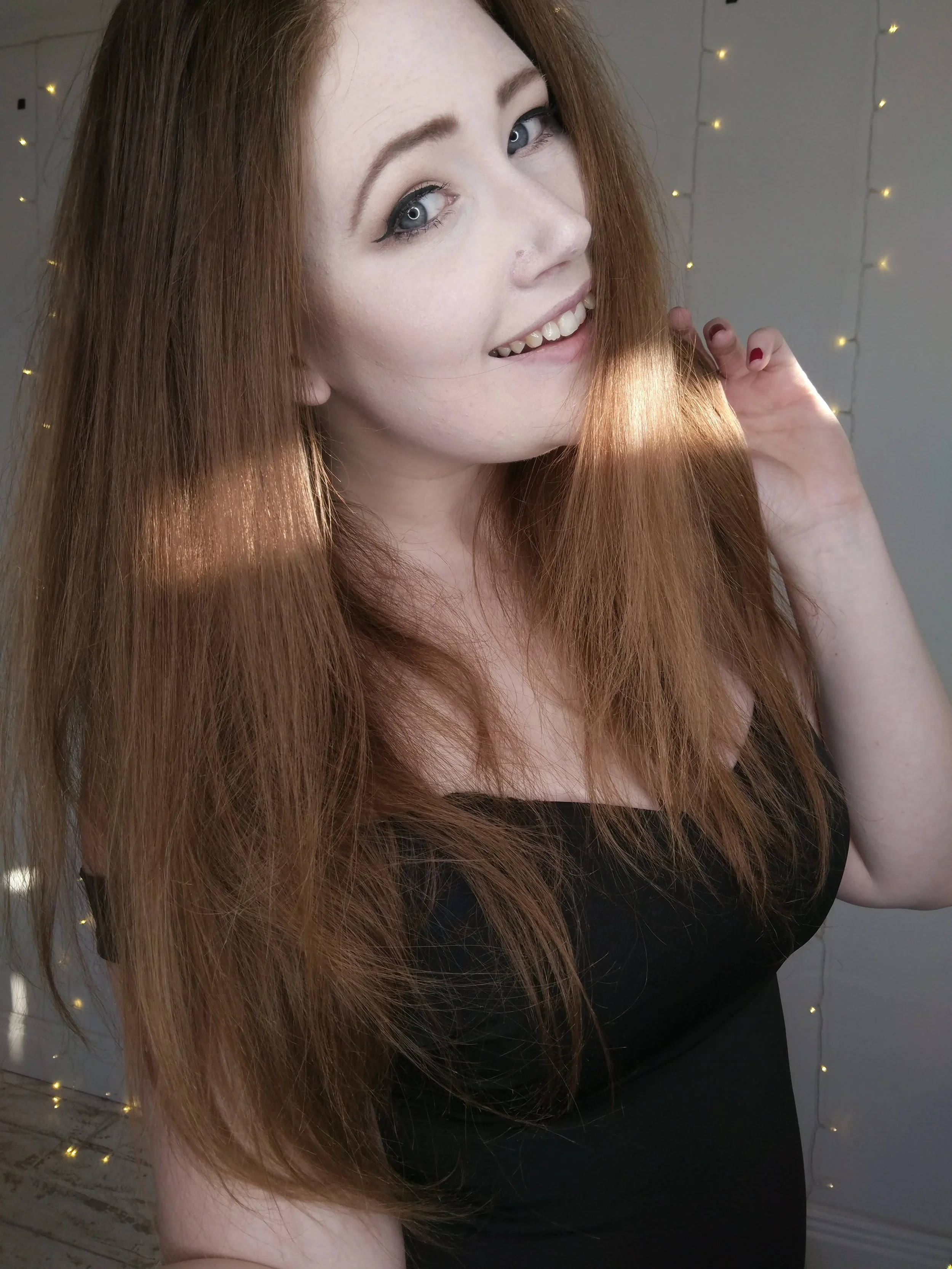 Close-up of a young woman with long red hair and gray eyes, smiling and touching her hair, set against a background of fairy lights.