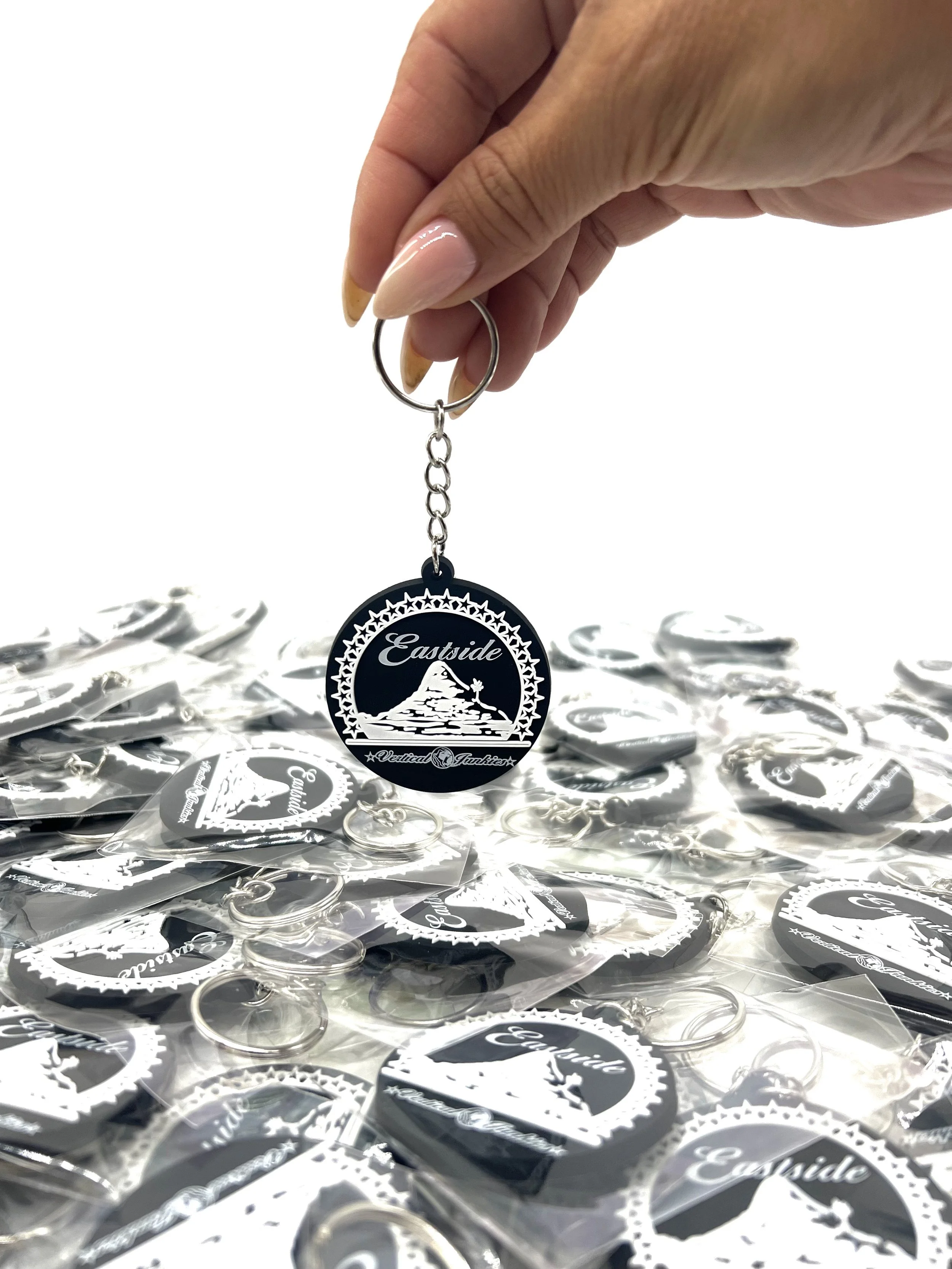 A hand holding a black keychain with a circular design that says 'Eastside' and features a mountain with a lake and trees, above the words 'Vertical Tourism'. Multiple similar keychains are laid out on a table below.