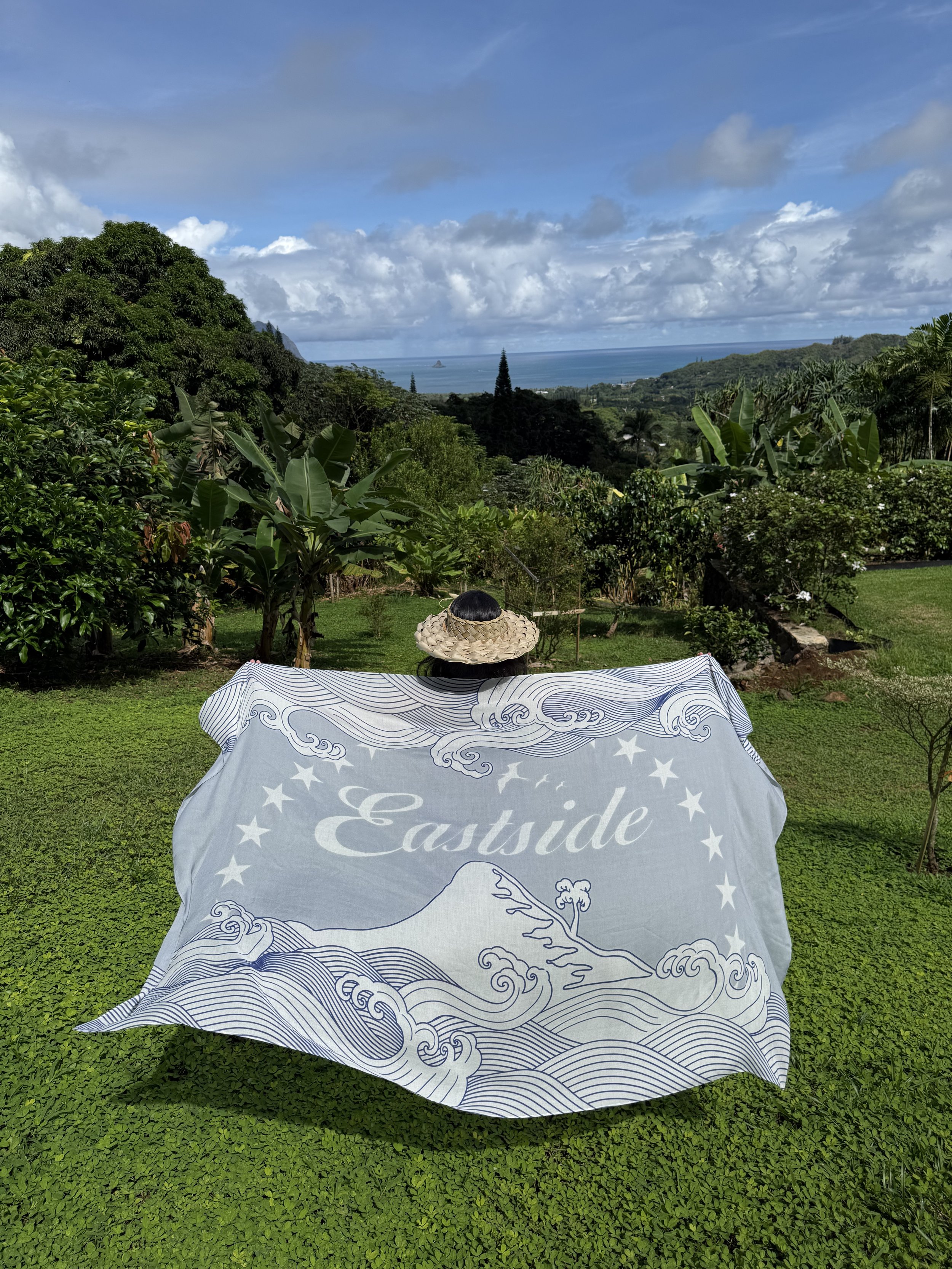 A person with a straw hat sitting on a lush green lawn, holding a large fabric with the word 'Eastside' and a wave design, overlooking a scenic view of trees, mountains, and the ocean under a blue sky with clouds.