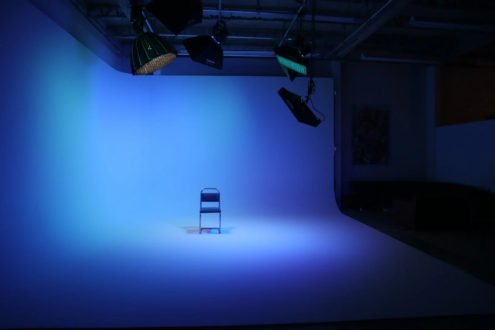 An empty chair on a photography studio backdrop illuminated with blue lighting, with professional studio lights hanging from the ceiling.