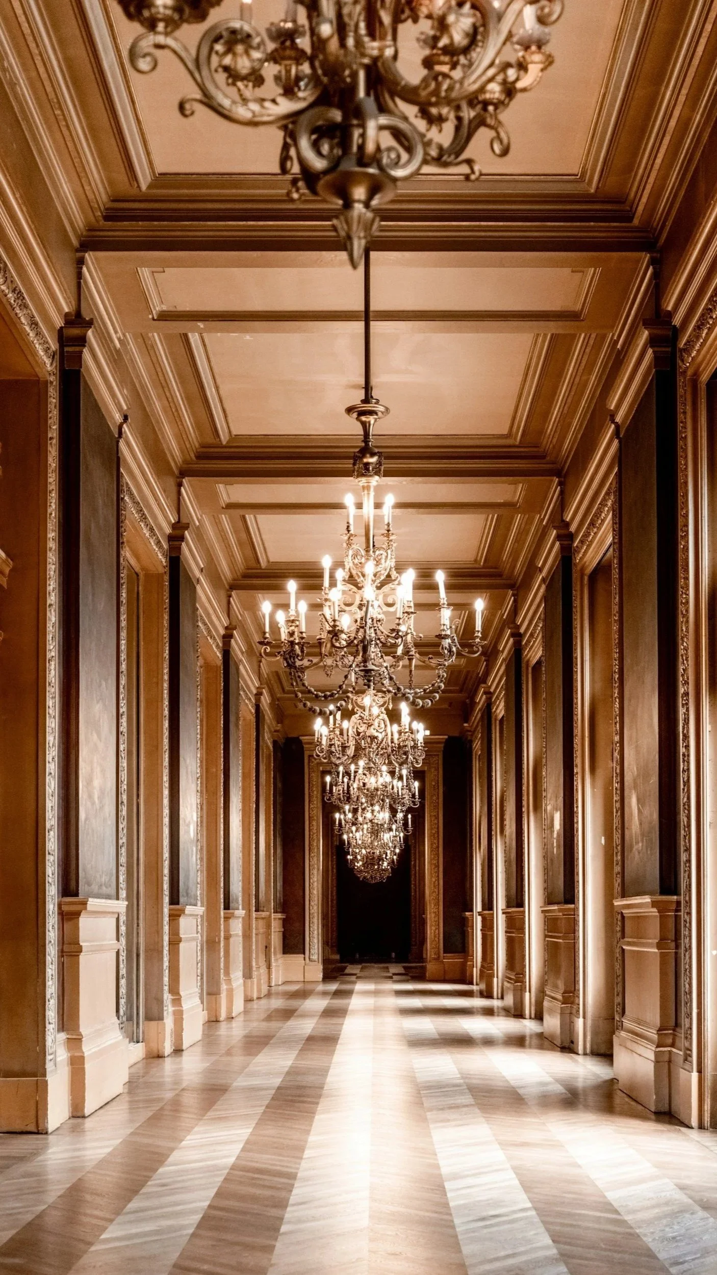 Elegant corridor with high ceilings, ornate crown molding, and multiple chandeliers hanging from the ceiling. The floor has a striped pattern of light and dark wood.