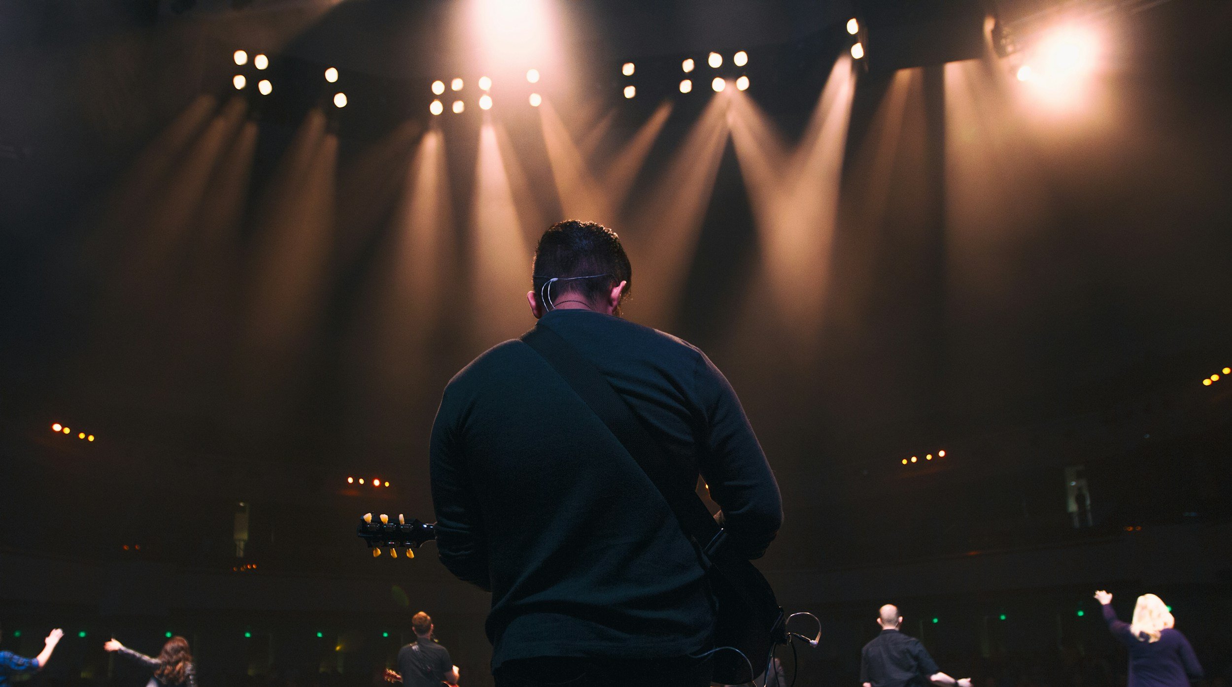 A musician on stage seen from the back, holding a guitar, with stage lights shining down from above, while a group of people in the audience raise their hands in excitement