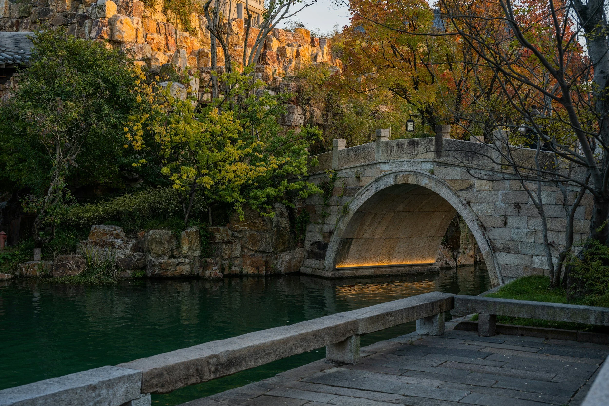 A stone bridge over a calm waterway, surrounded by green and autumn-colored trees, with rocks and a rocky hillside in the background.