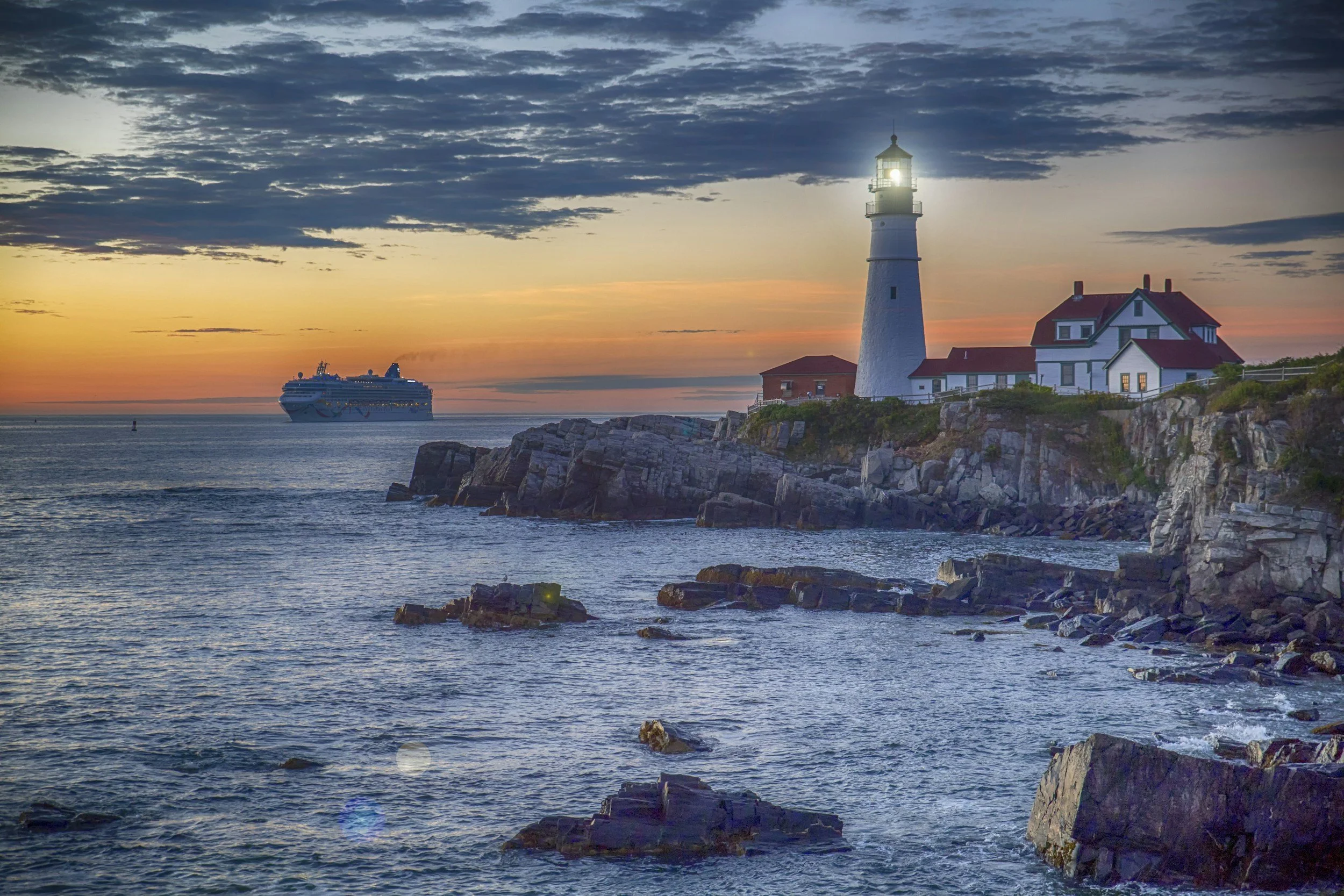 A lighthouse on a rocky coastline at sunset with a large cruise ship in the distance and a partly cloudy sky.