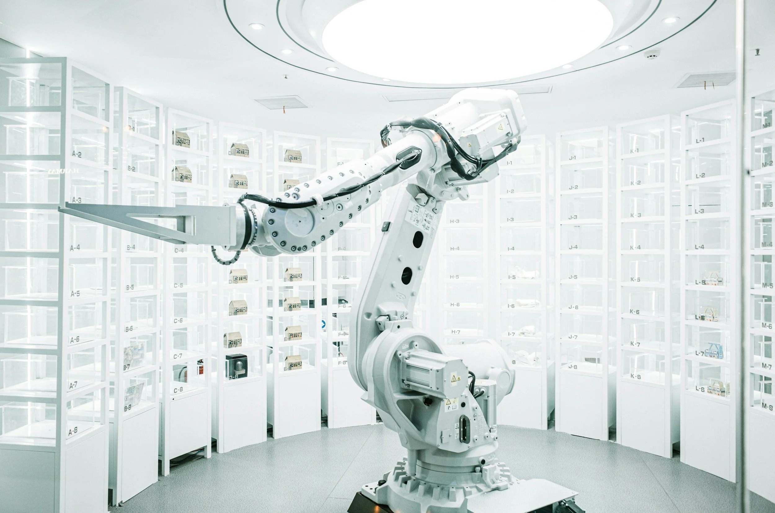 Robotic arm inside a sanitary, high-tech storage facility or laboratory, surrounded by numerous white shelves filled with small boxes or equipment.