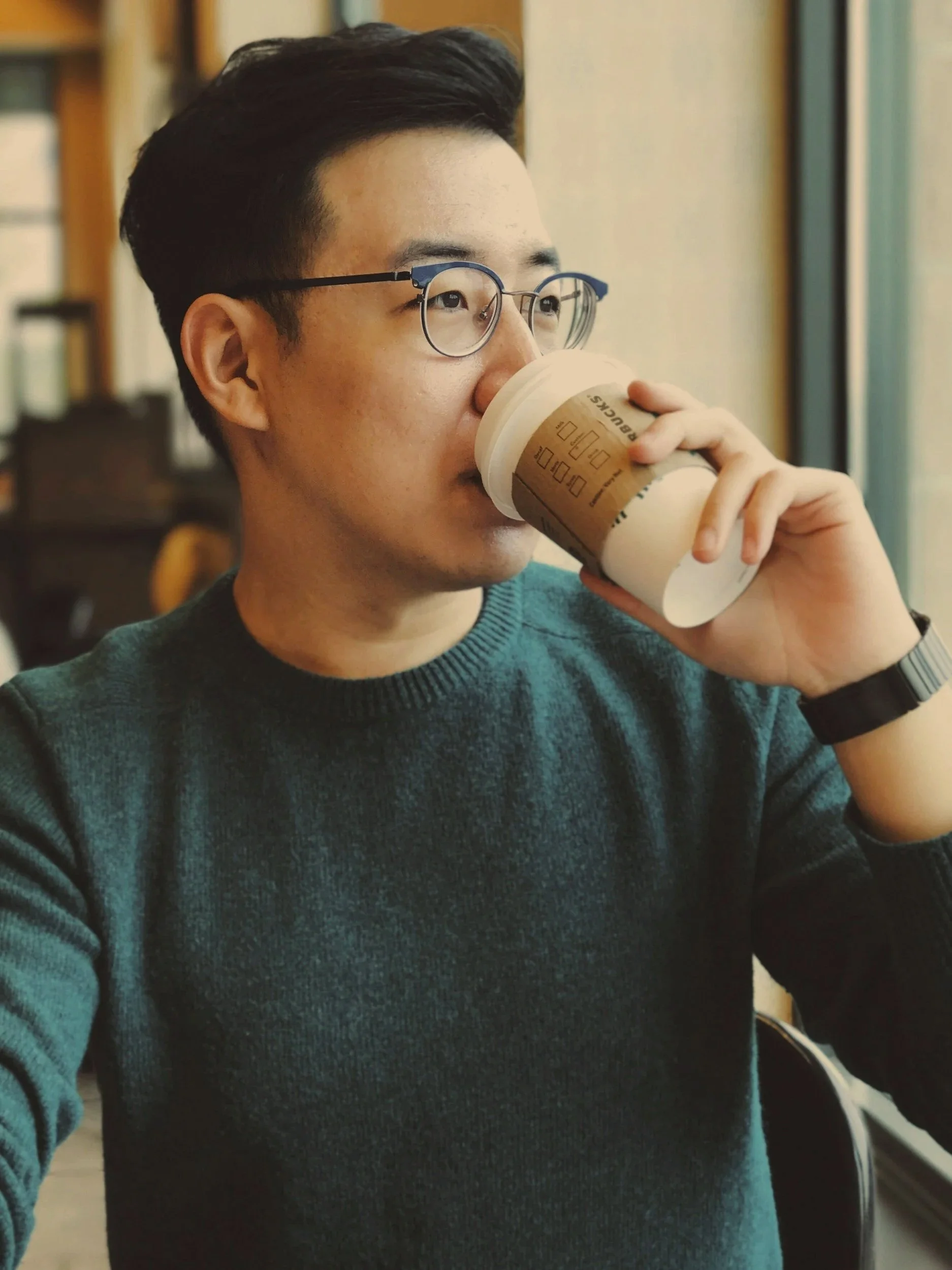 A man wearing glasses and a green sweater drinks coffee from a paper cup while sitting by a window.